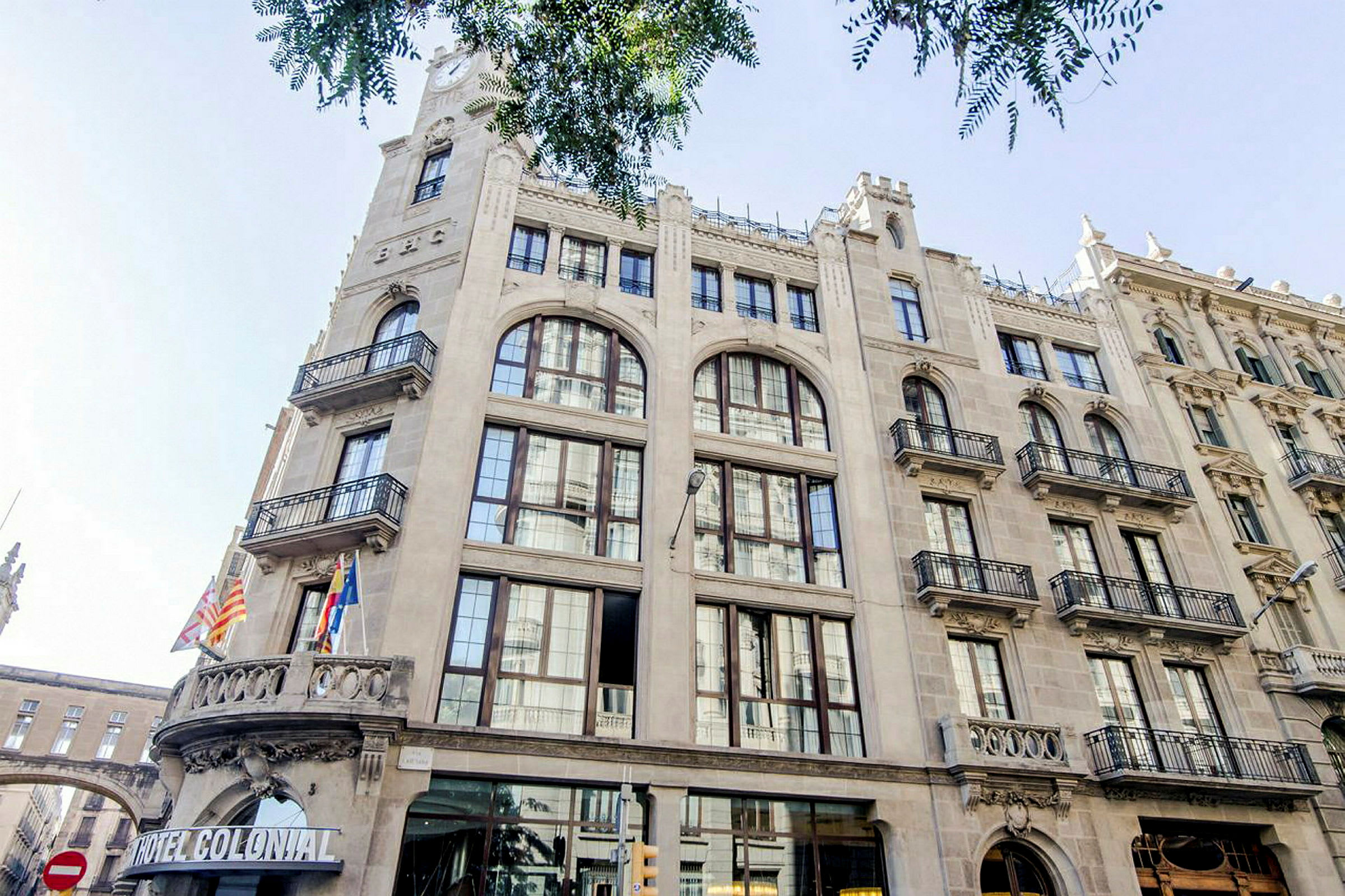Byggnad i klassisk arkitektur med stora fönster och balkonger prydd av flaggor vid entrén, inkluderande skylt med texten 'Hotel Colonial'. (AI-genererad text)