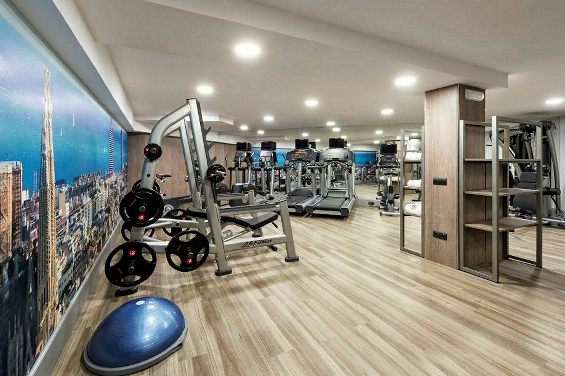 Ett gym med träningsutrustning och maskiner, inklusive löpband, vikter och cyklar, på ett ljust trämönstrat golv. (AI-genererad text)