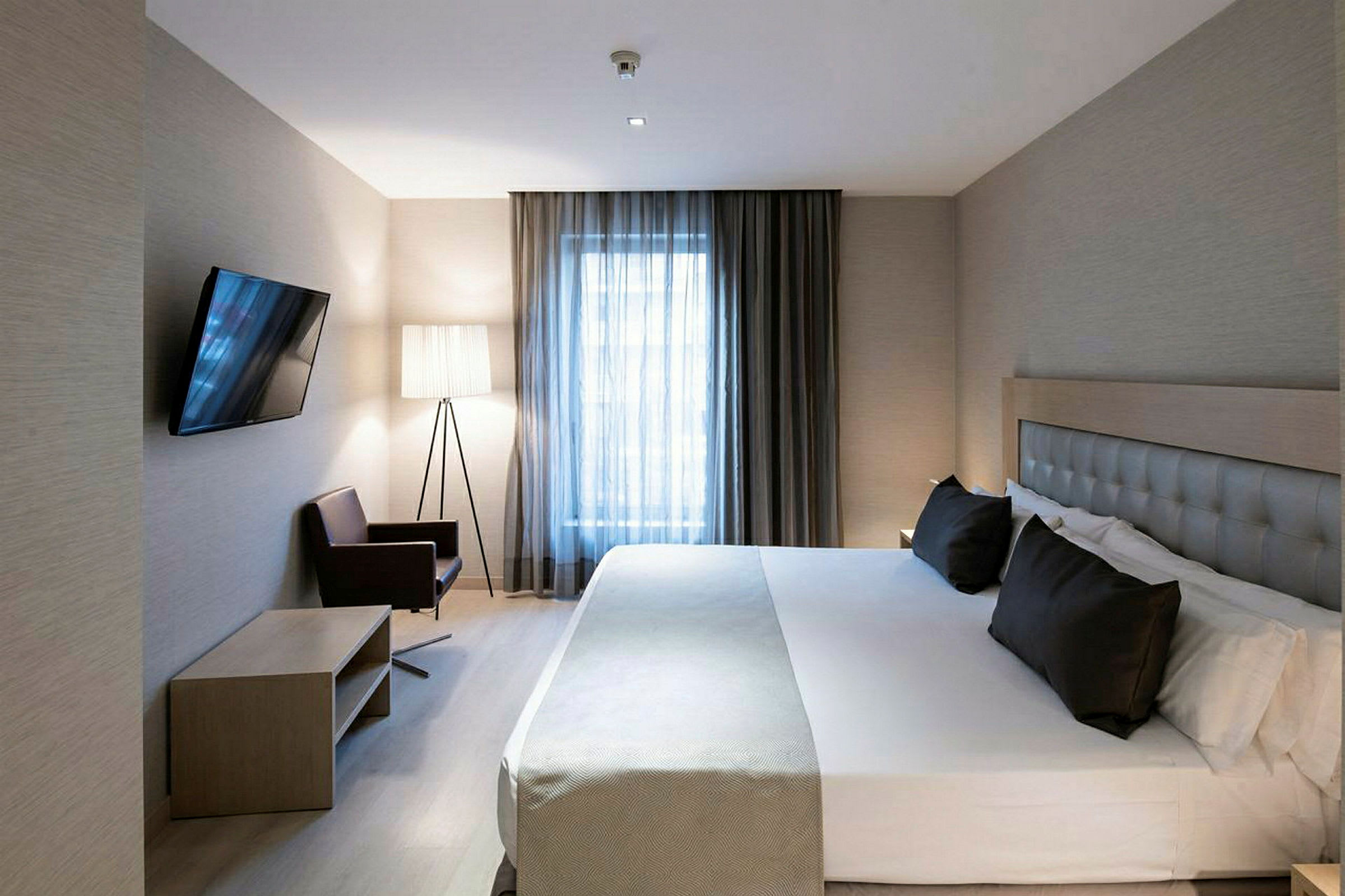 Hotellrum med en stor säng, vägg-TV, fåtölj och ett fönster med gardiner. (AI-genererad text)