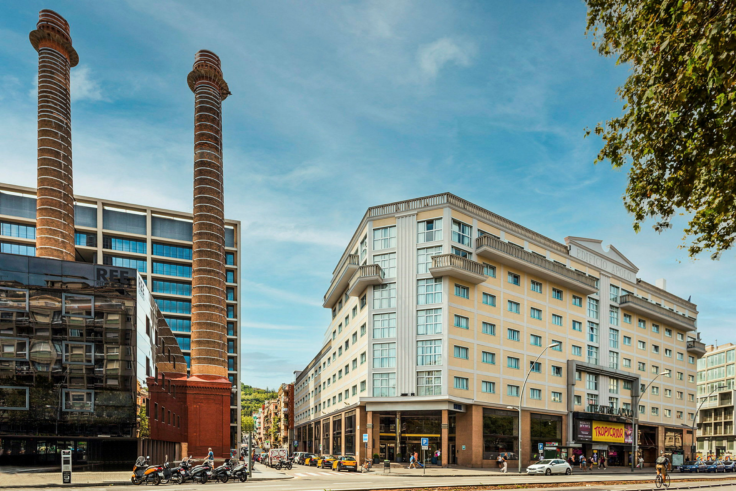 Bild av en stadsmiljö med moderna byggnader, två höga skorstenar och några människor och fordon i förgrunden. (AI-genererad text)