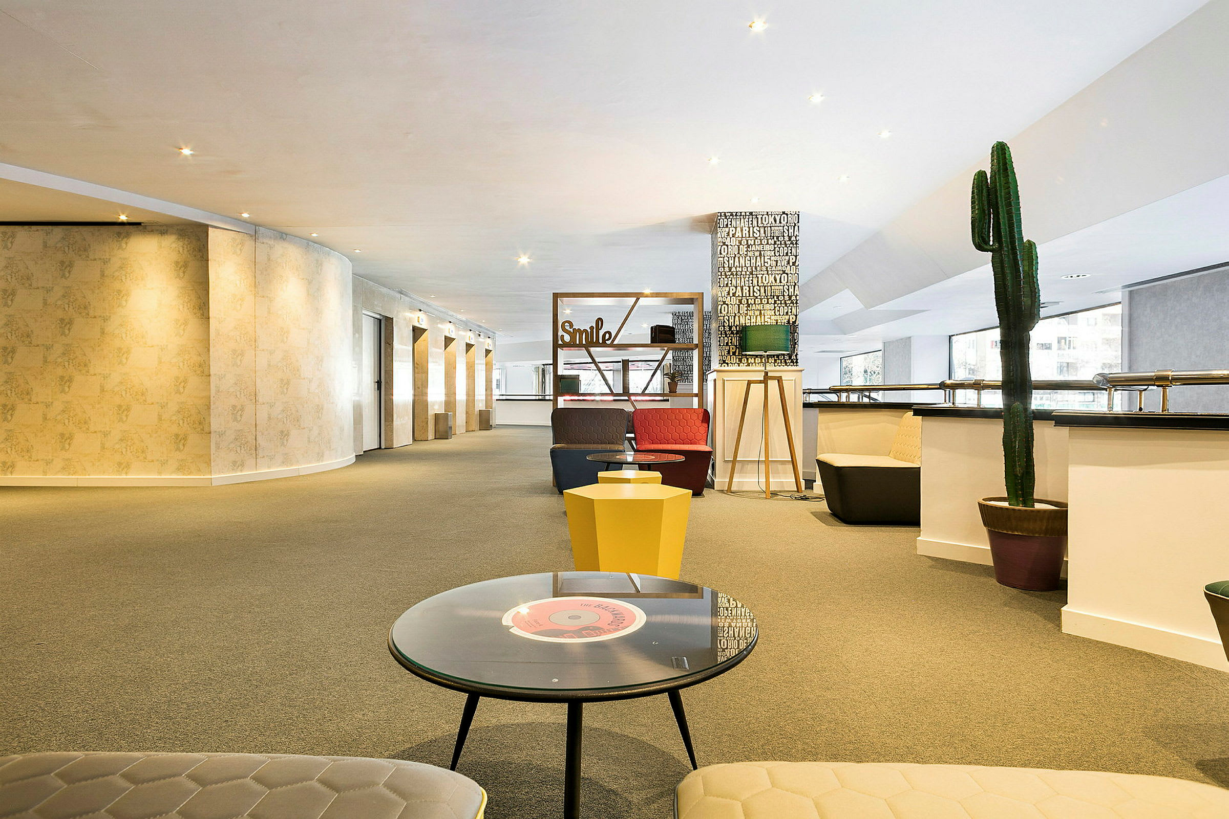 En rymlig och ljus lounge med sittplatser, bord och dekorationer inklusive en stor kaktus och skyltar med text. (AI-genererad text)