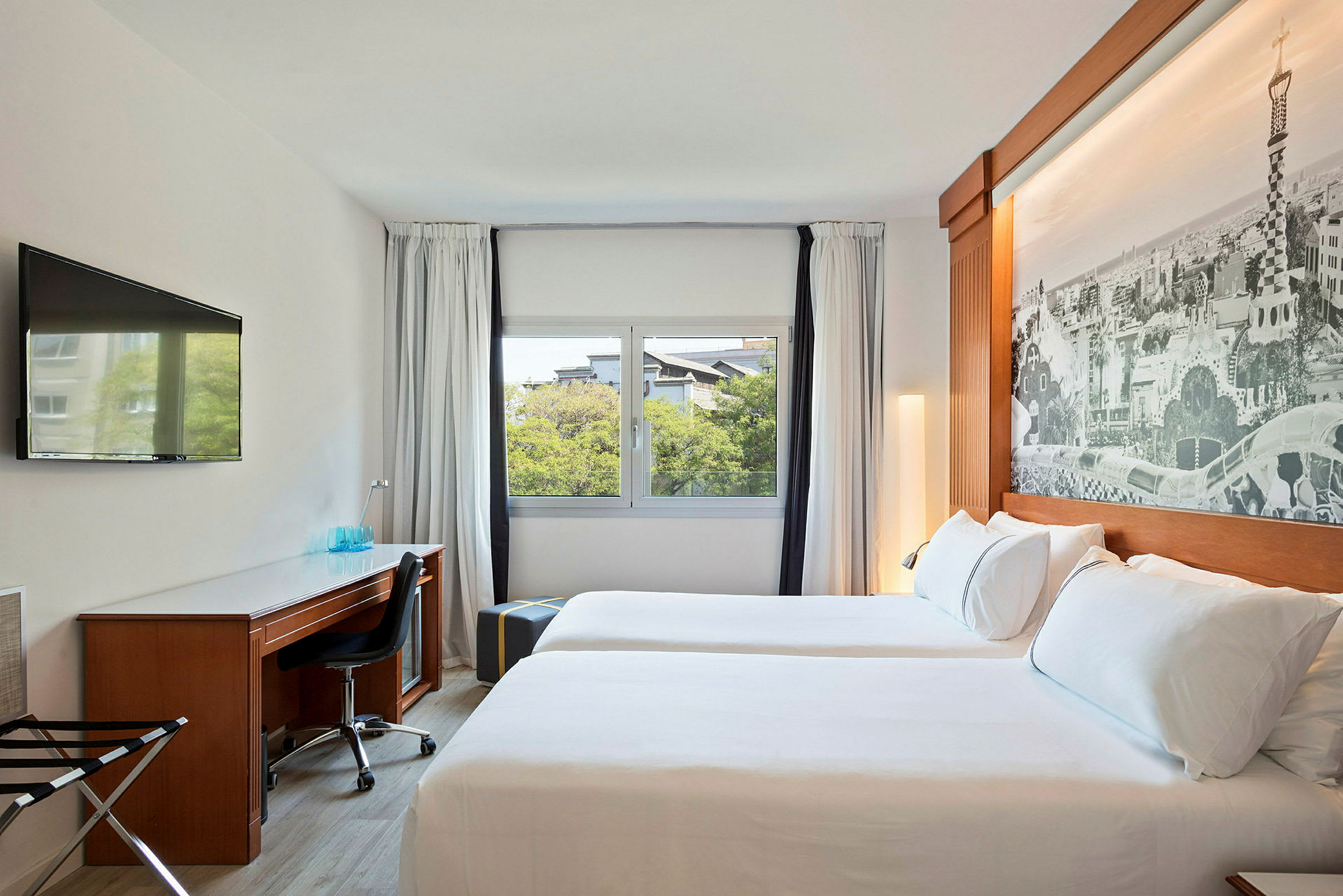 Ett hotellrum med två sängar, ett skrivbord med stol, en väggmonterad TV och ett stort fönster med utsikt. (AI-genererad text)