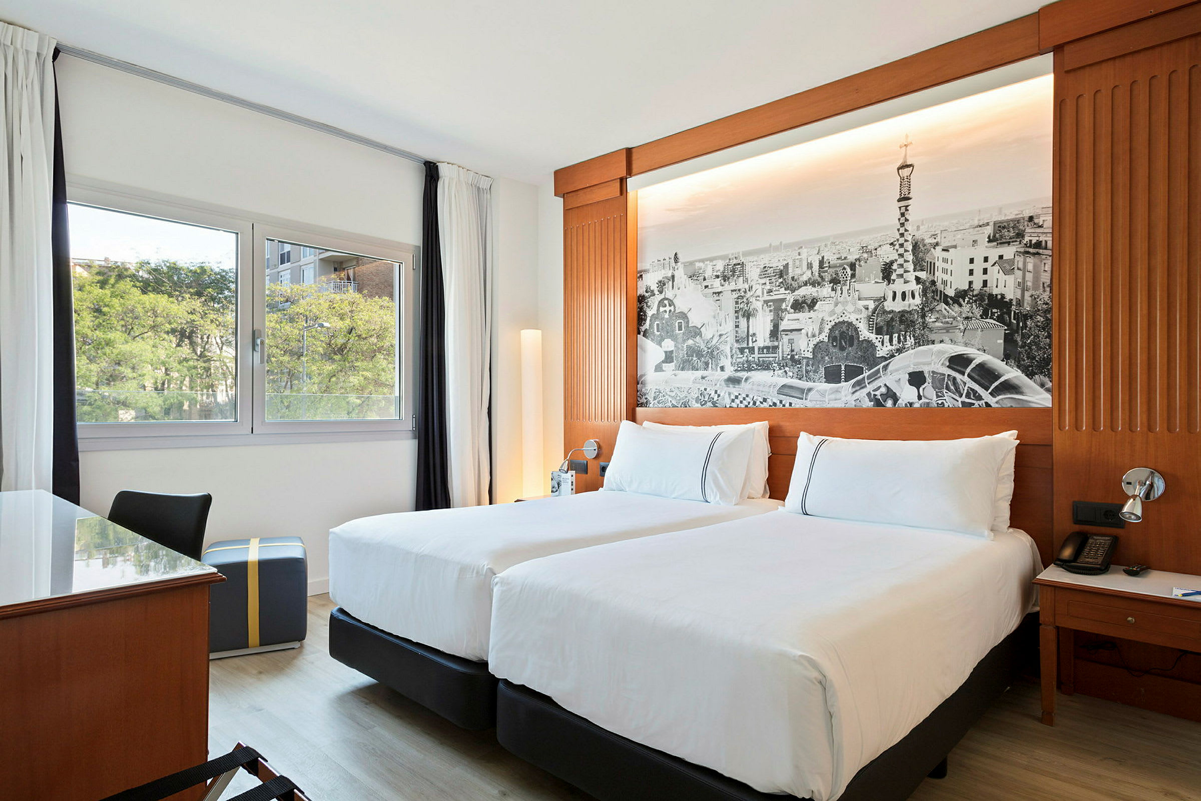 Ett hotellrum med två enkelsängar, ett skrivbord, en fönstervy mot grönska och en vägg med en stor svartvit bild som dekor. (AI-genererad text)