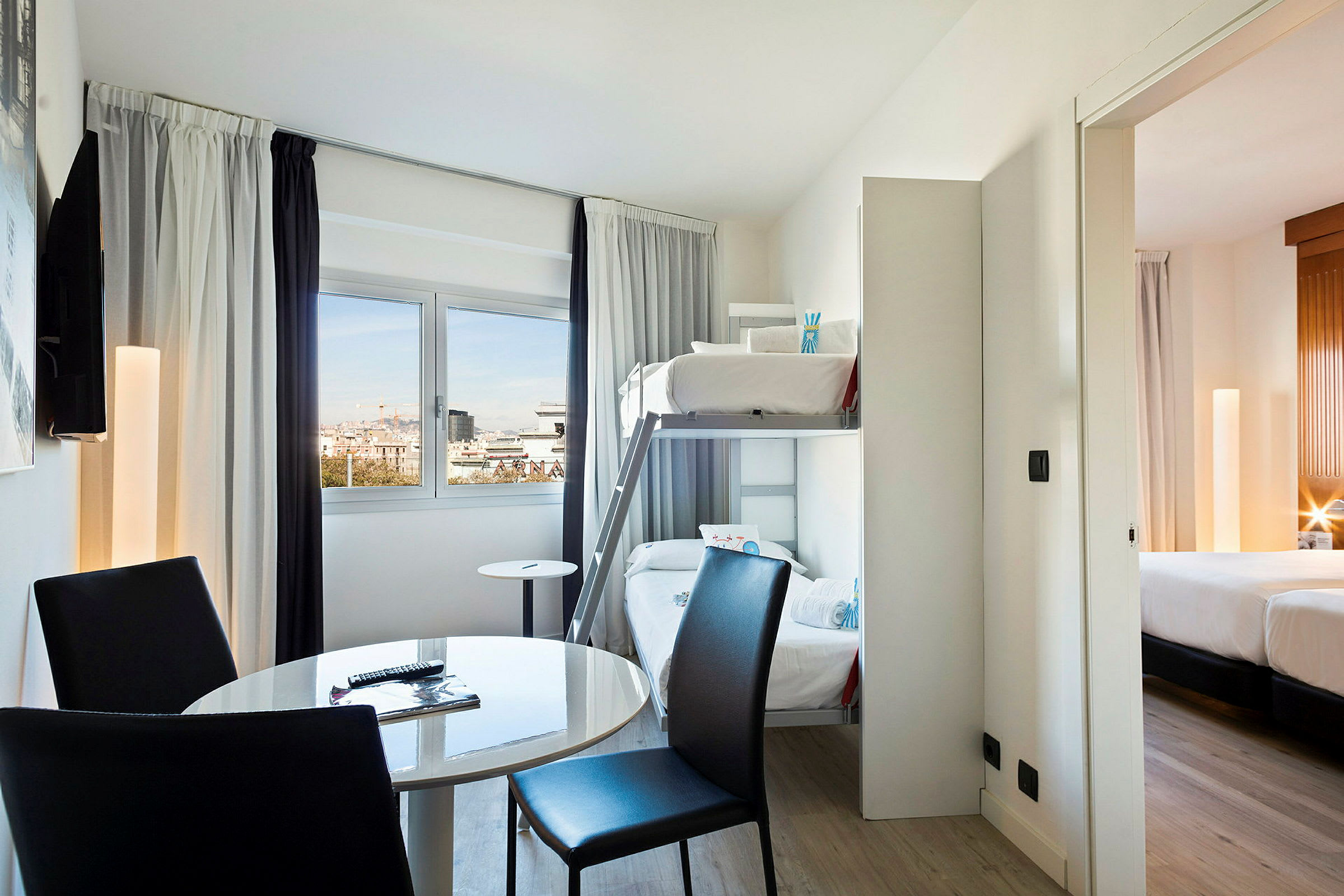 Ett hotellrum med våningssängar, ett runt bord och stolar, samt en vy genom fönstret. Ett anslutande sovrum med dubbelsäng syns. (AI-genererad text)