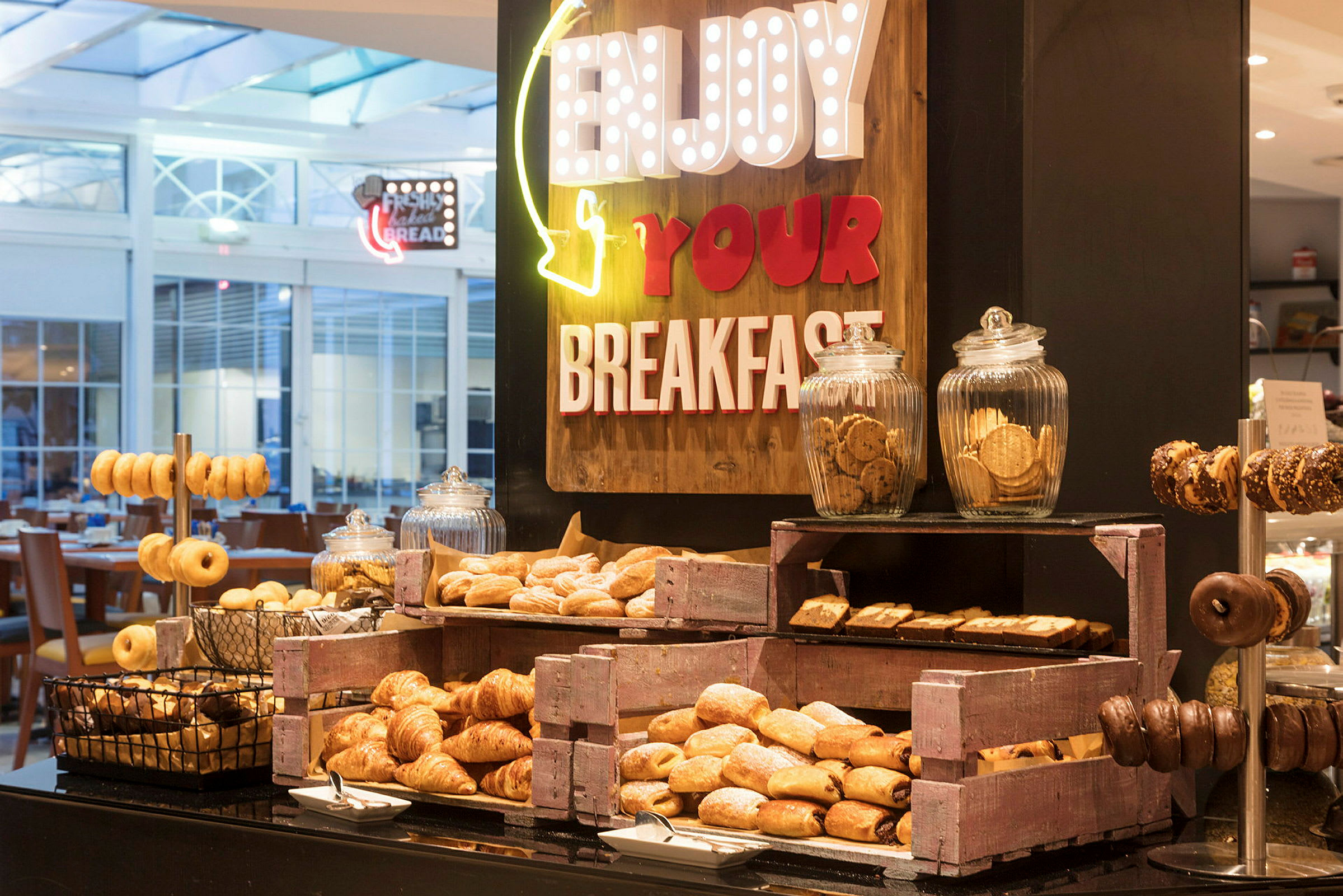 En buffé med olika frukostbakverk såsom croissanter, munkar och småkakor, arrangerad med skyltar som uppmuntrar till att njuta av frukosten. (AI-genererad text)