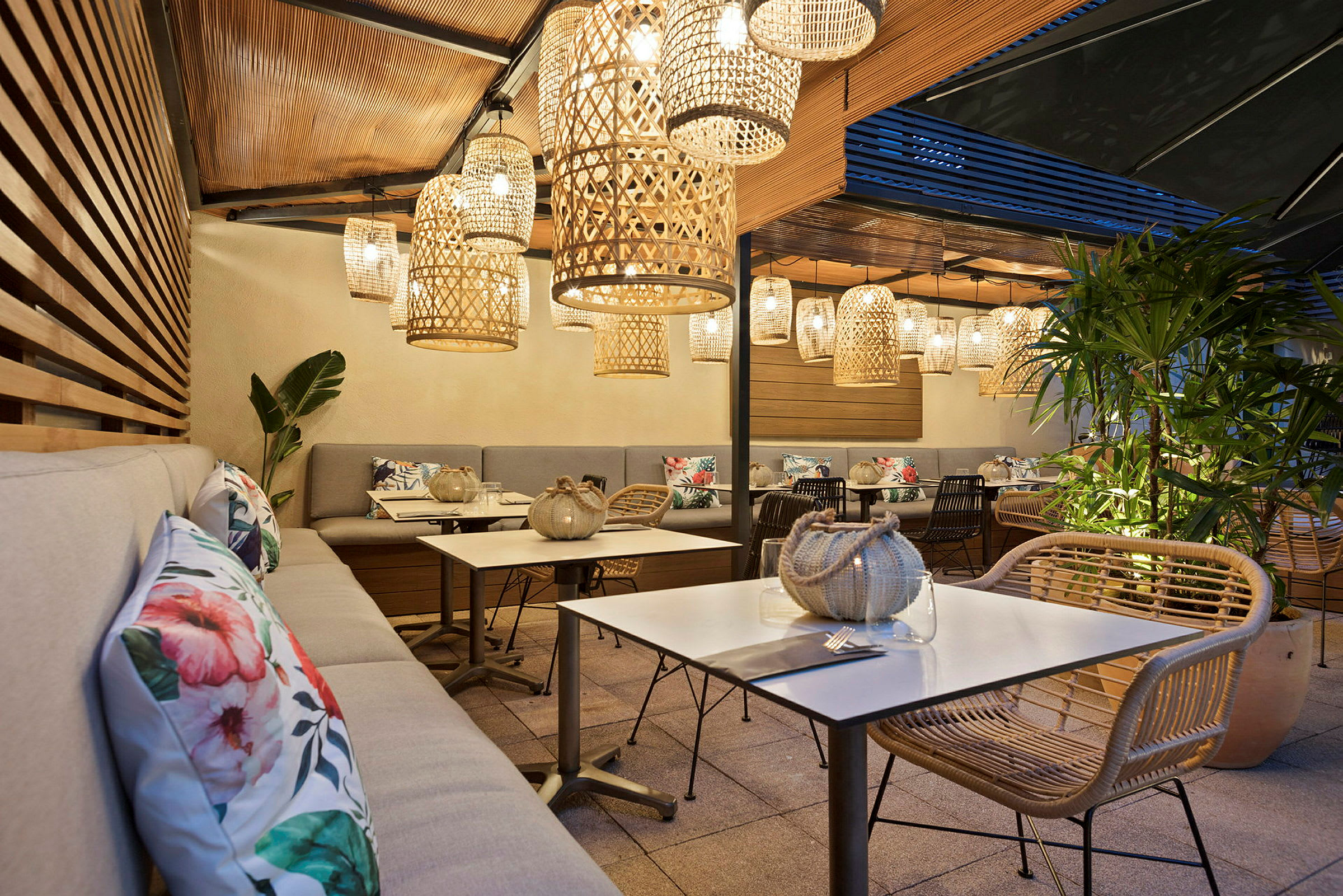 Terrass med sittplatser, bord och dekorativa korglampor som hänger från taket. Växter och prydnadskuddar synliga. (AI-genererad text)