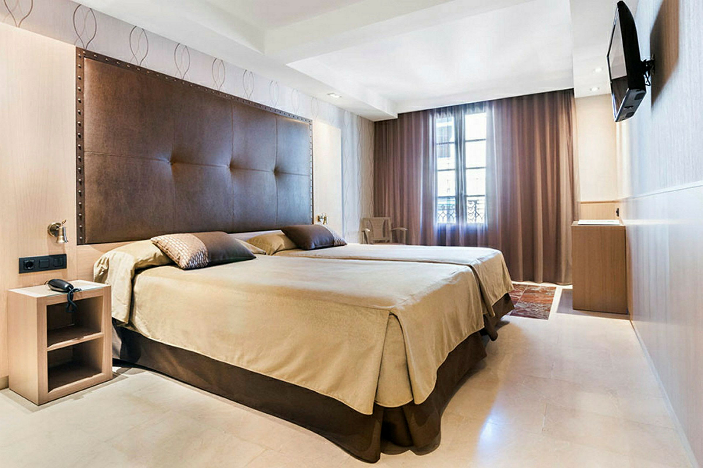 Ett hotellrum med en stor säng, brun sänggavel och beige sängkläder. En TV är monterad på väggen och fönstret är täckt av mörka gardiner. (AI-genererad text)