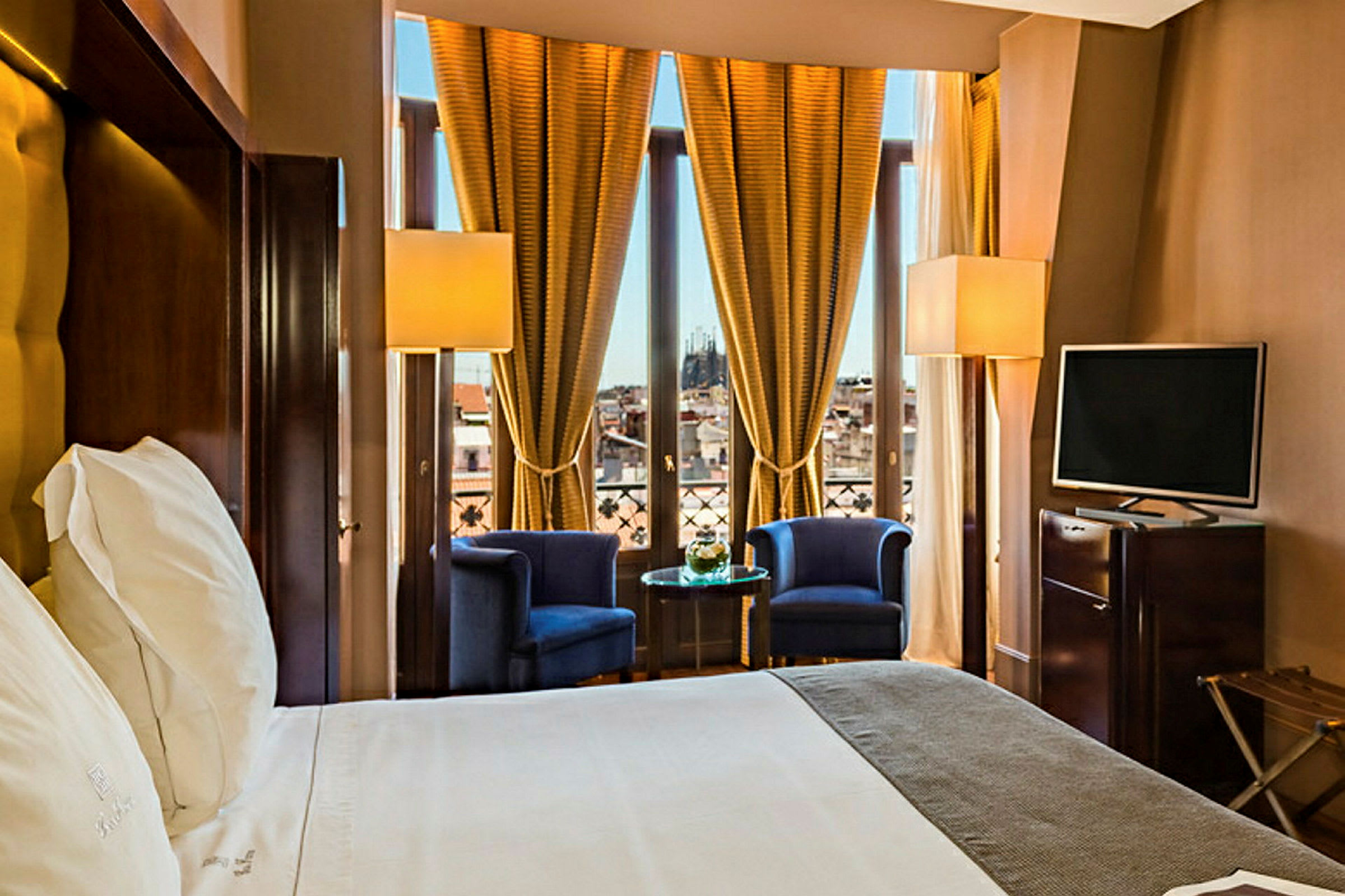 Ett hotellrum med en stor säng, två fåtöljer och ett sidobord framför en fönstervägg med gyllene gardiner. (AI-genererad text)