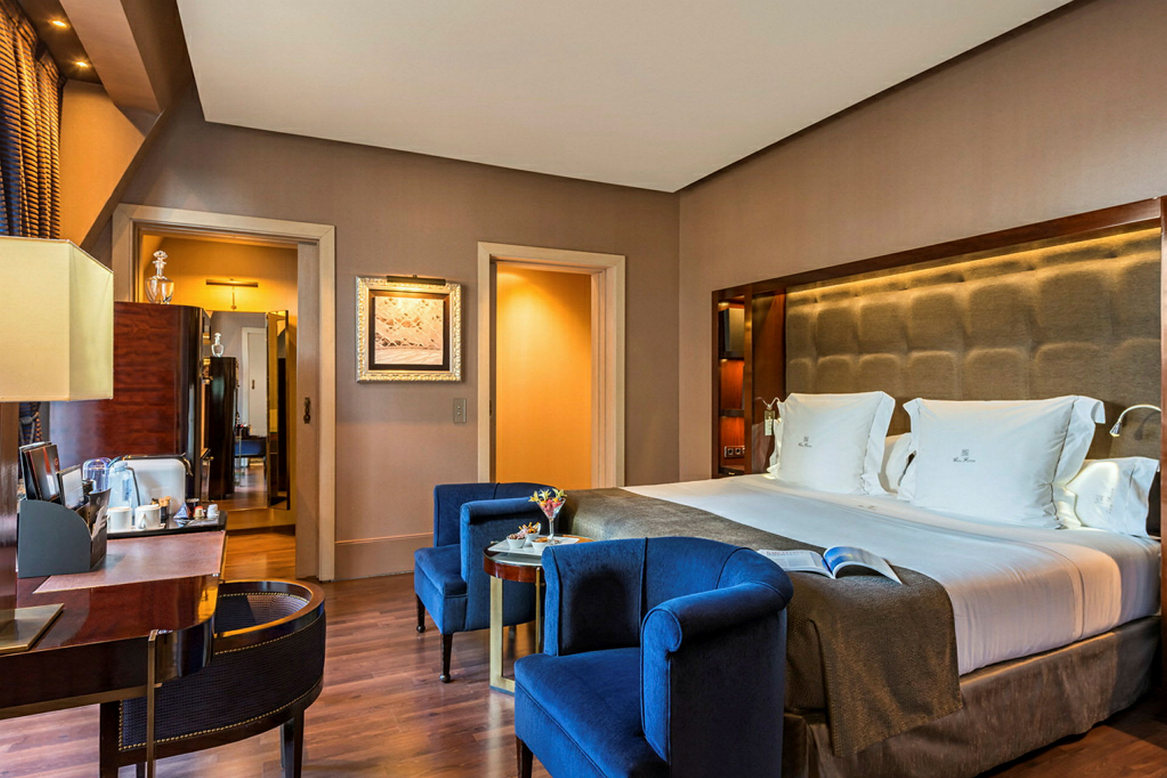 Ett elegant hotellrum med en dubbelsäng och blå fåtöljer framför ett bord. (AI-genererad text)