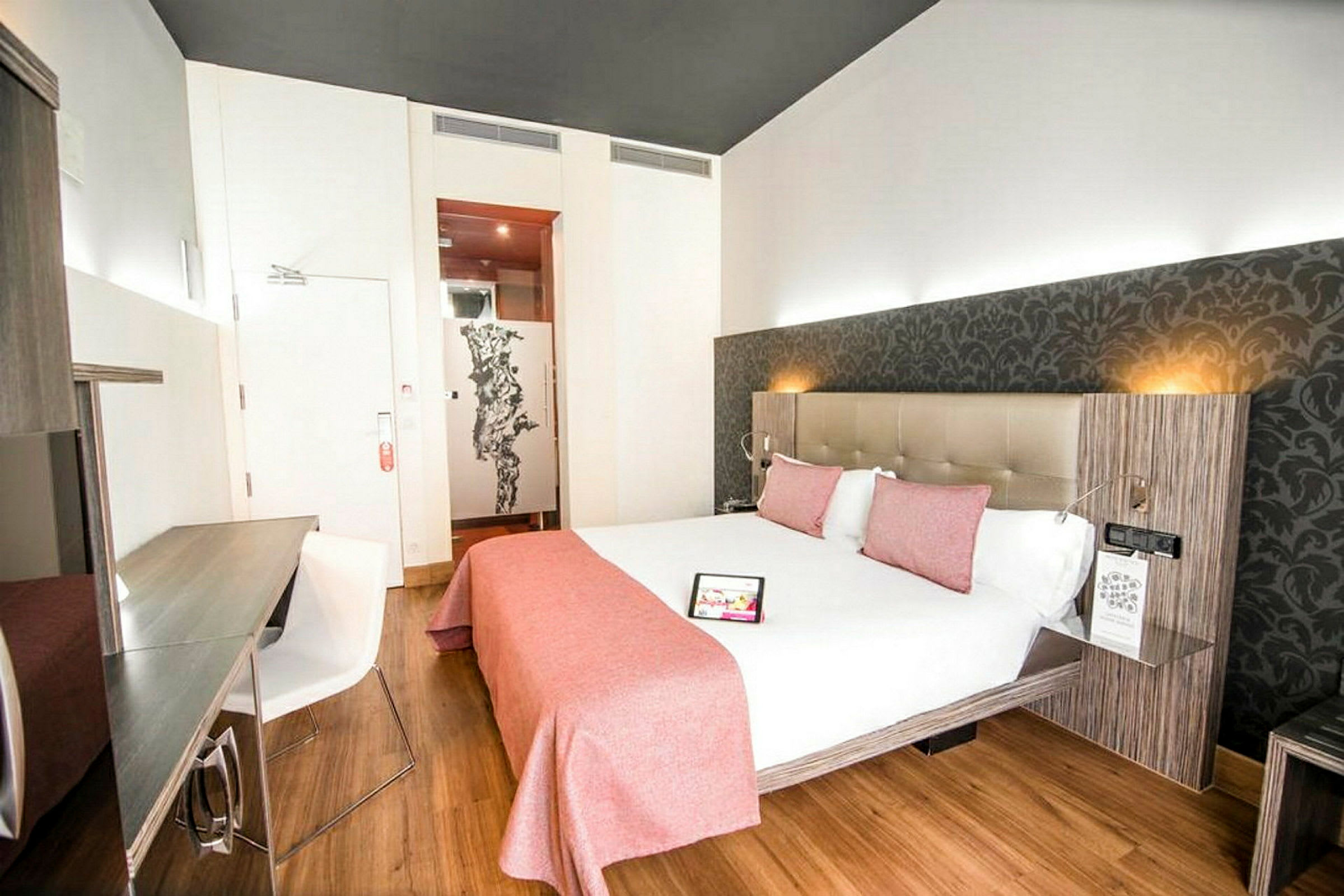 Ett ljust hotellrum med en dubbelsäng bäddad med vita lakan och rosa sängkläder, ett skrivbord med en stol och en dekorativ vägg bakom sängen. (AI-genererad text)