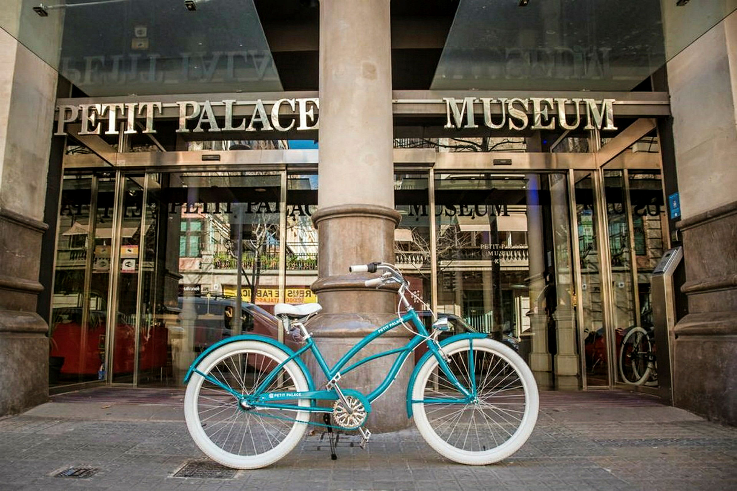 En turkos cykel med vita däck är parkerad framför entrén till ett hotell med texten 'Petit Palace Museum'. (AI-genererad text)