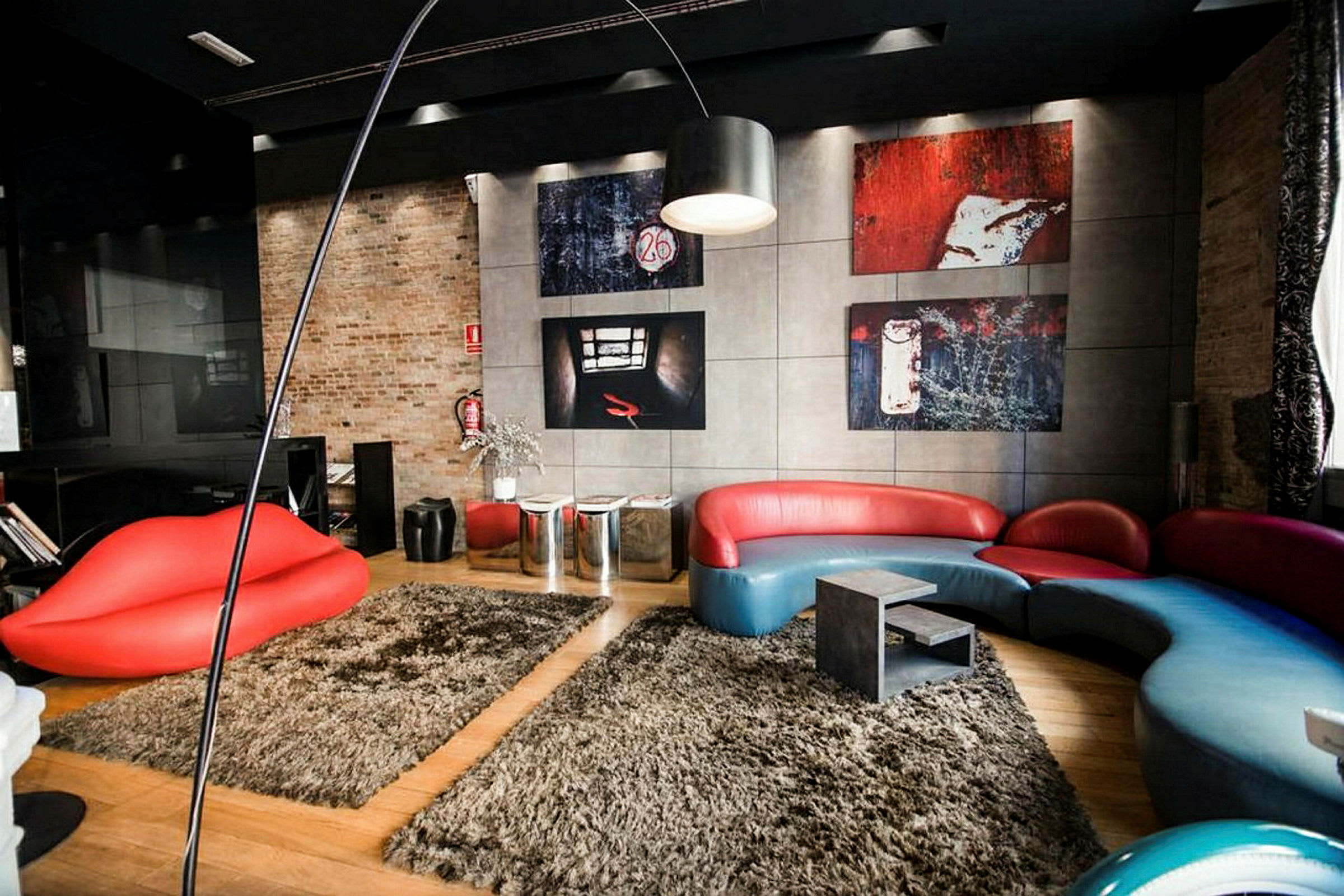 Ett modernt rum med en röd och blå soffa i unik design, en röd soffliknande sittmöbel formad som läppar, och konstverk på väggarna. Golvet är täckt av beige mattor. (AI-genererad text)