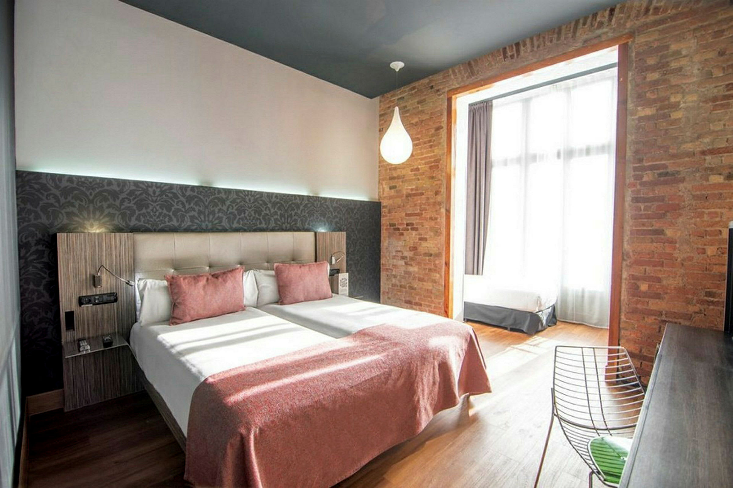 Ett hotellrum med två enkelsängar, rosa överkast och en tegelvägg vid ett stort fönster. (AI-genererad text)