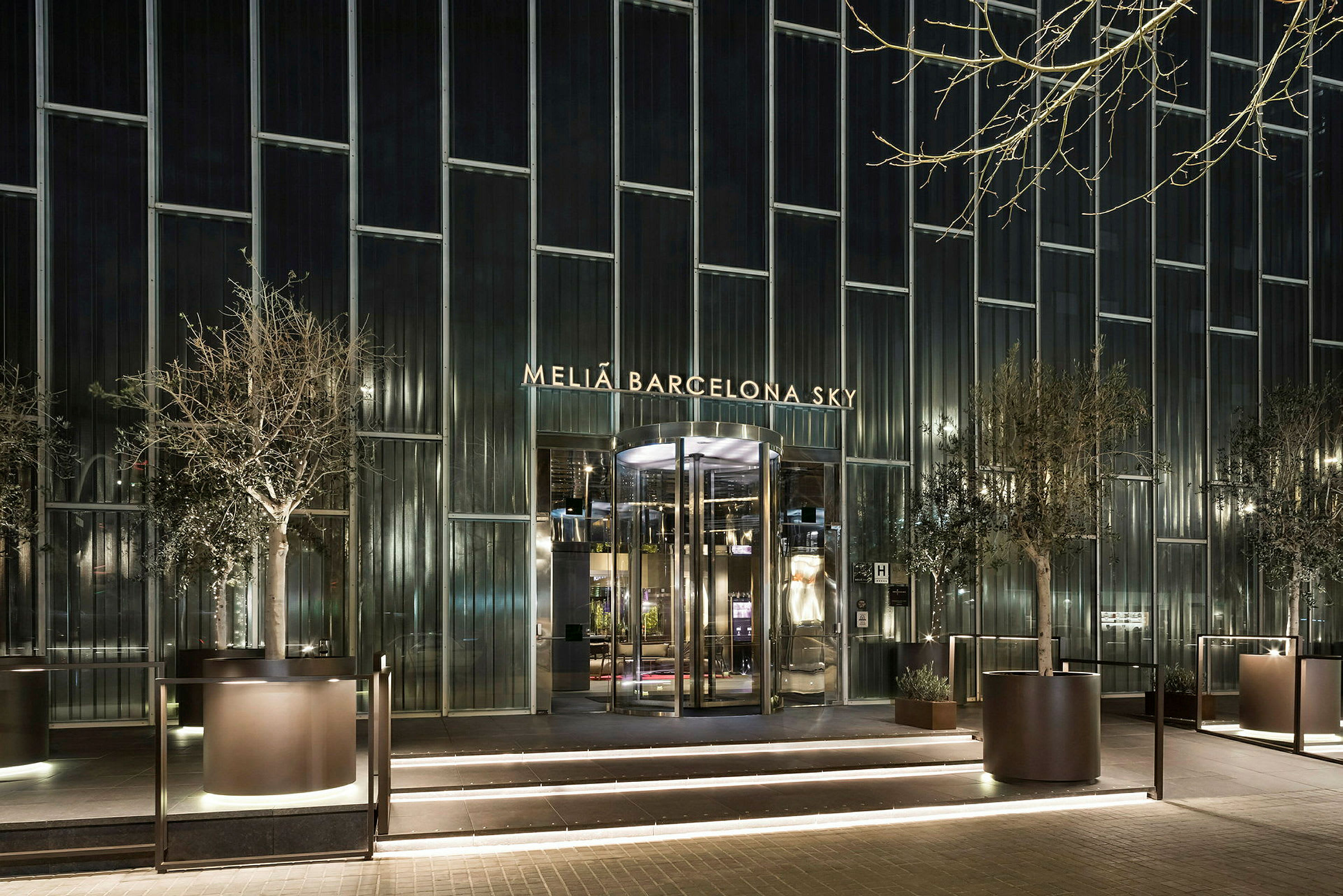 Fasaden av Melia Barcelona Sky Hotel med belyst entré och stora glasfönster, omgiven av planterade träd. (AI-genererad text)