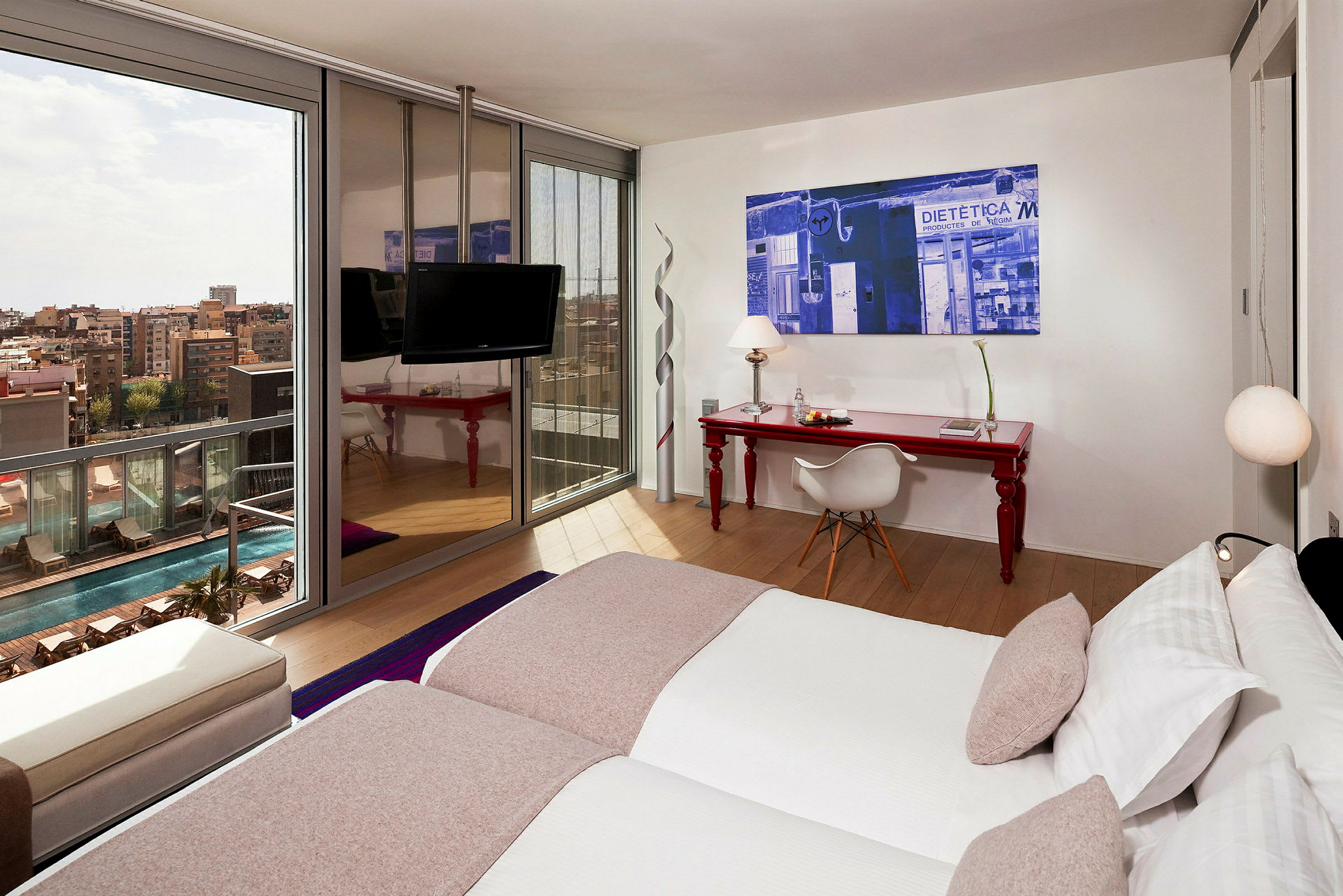 Ett modernt hotellrum med två enkelsängar, en röd skrivbord med stol, och ett stort fönster som vetter mot poolområdet och staden. (AI-genererad text)
