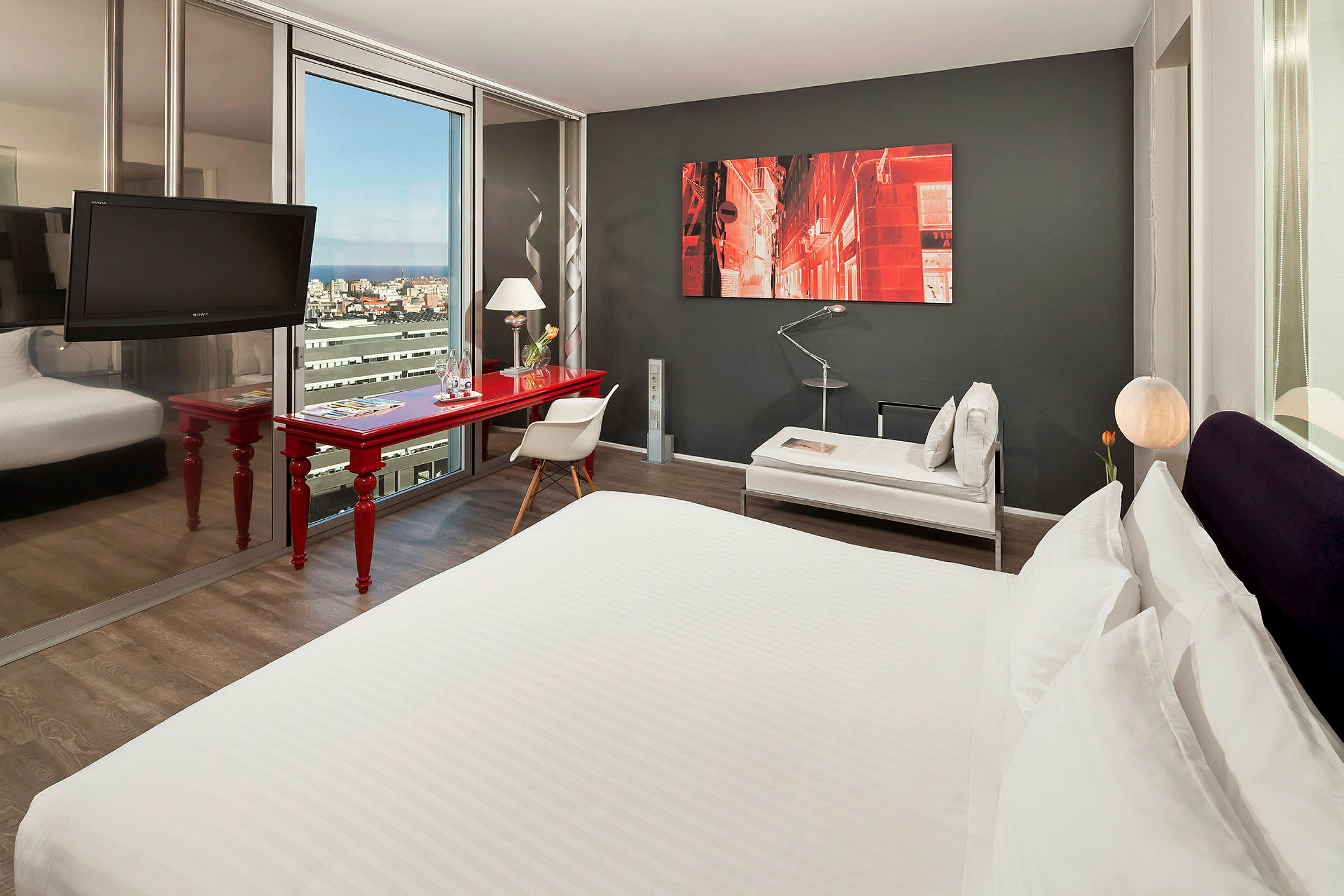 Ett modernt hotellrum med en stor säng, en röd skrivbord, en stol och ett soffbord. En målning pryder den mörkgrå väggen, och ett stort fönster visar en stadsutsikt. (AI-genererad text)