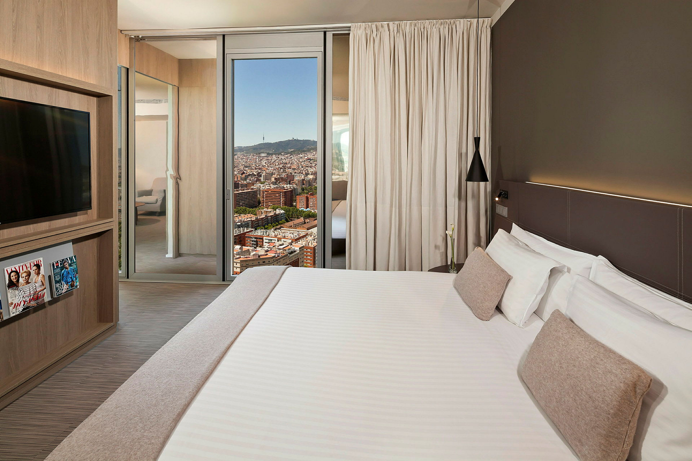 Ett hotellrum med en stor säng med vita sängkläder och beiga kuddar, en modern design med ett TV-skåp, tidskrifter och ett fönster som visar en stadsvy. (AI-genererad text)