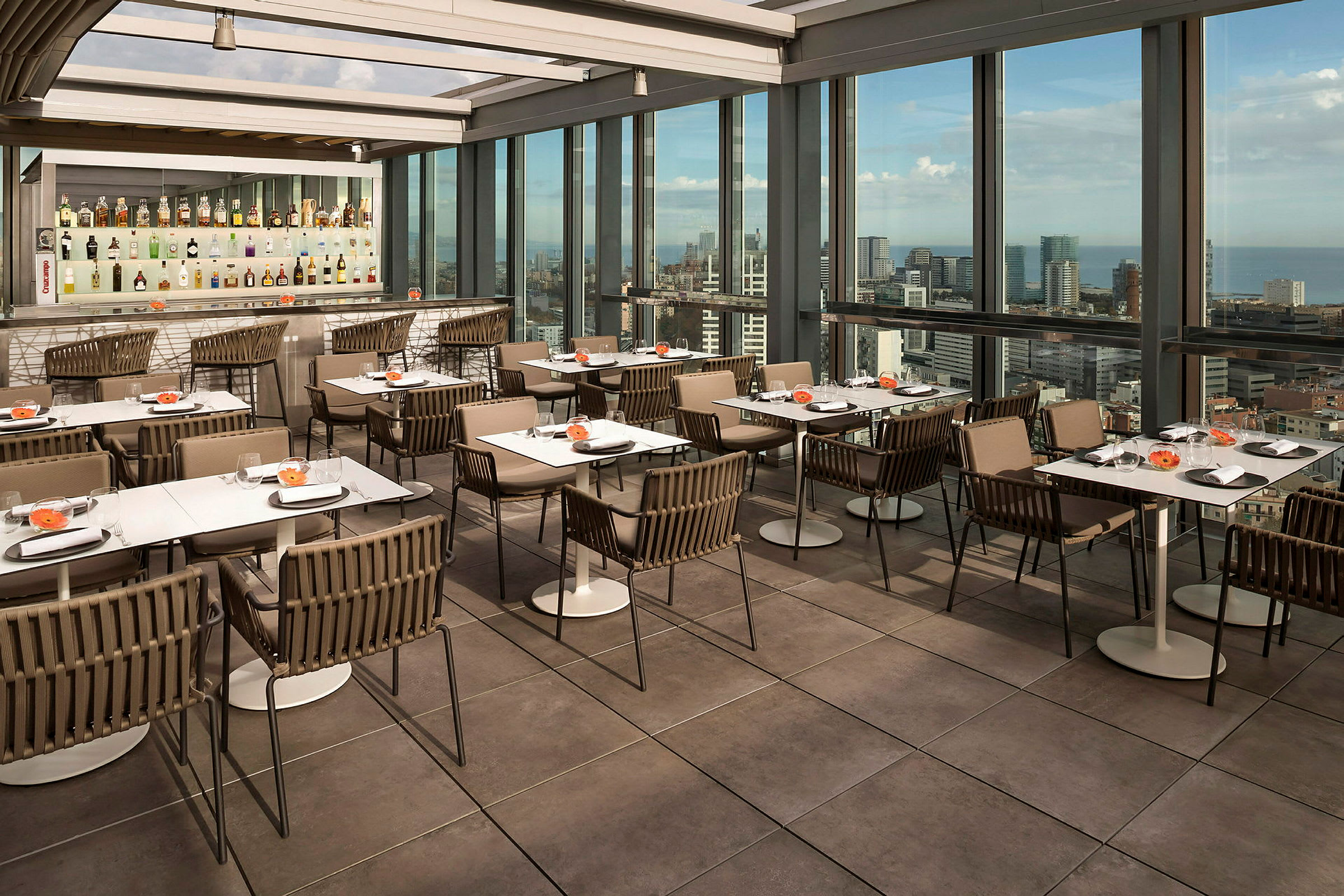 En restaurang med bord och stolar på en terrass med stora glasfönster som visar en stadsutsikt. (AI-genererad text)