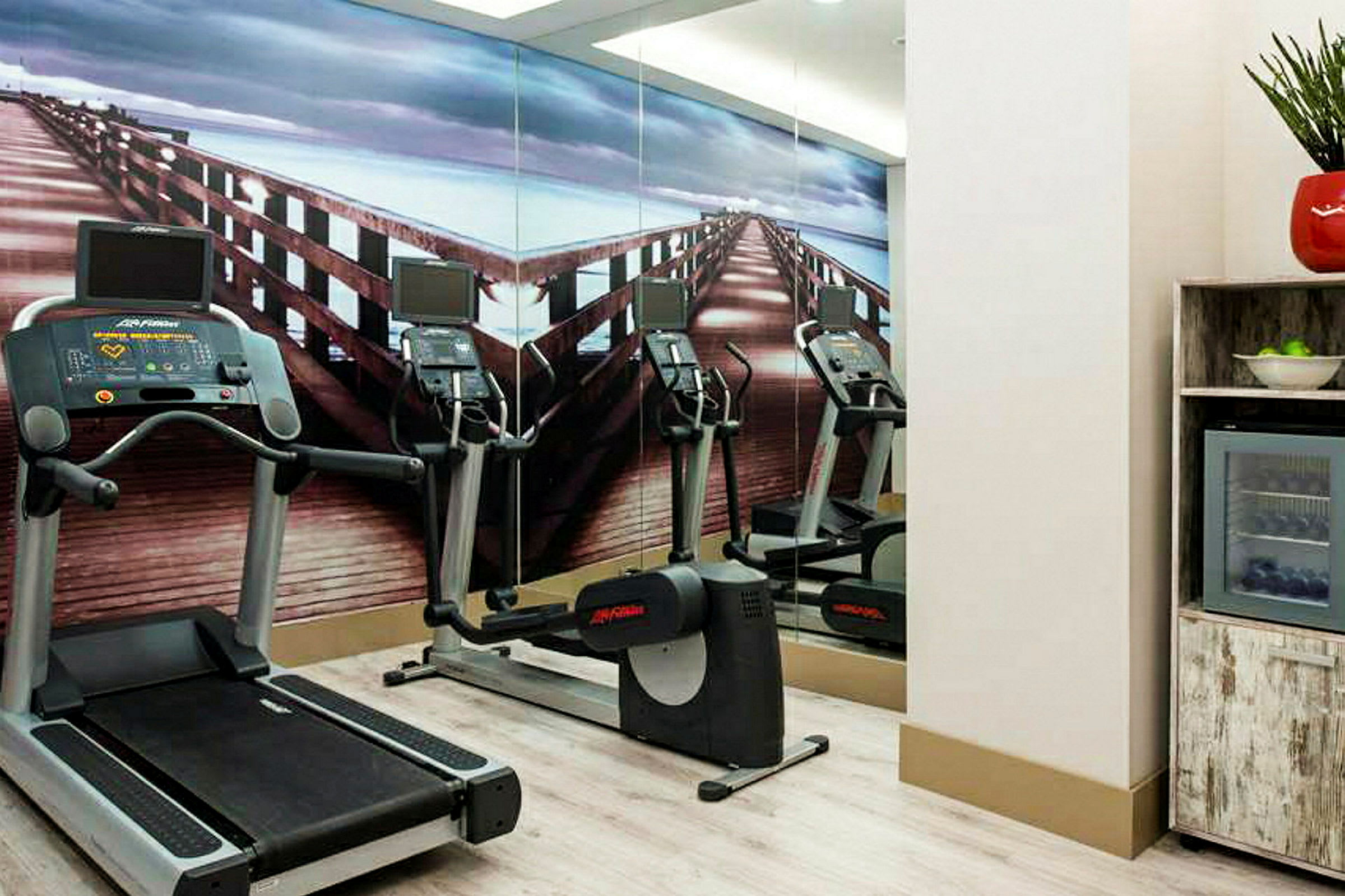 Ett gym med motionsutrustning och en vägg med en bild av en träbrygga. (AI-genererad text)