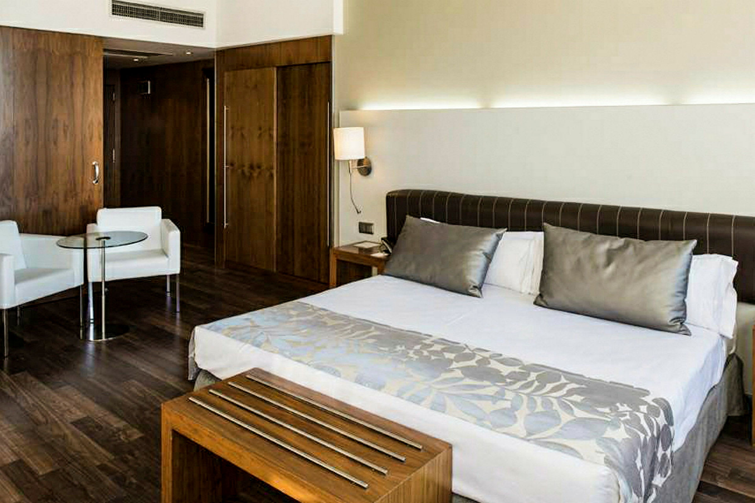 Ett hotellrum med en stor säng, två vita fåtöljer och ett glasbord. Rummet har träbeklädda väggar och modern inredning. (AI-genererad text)