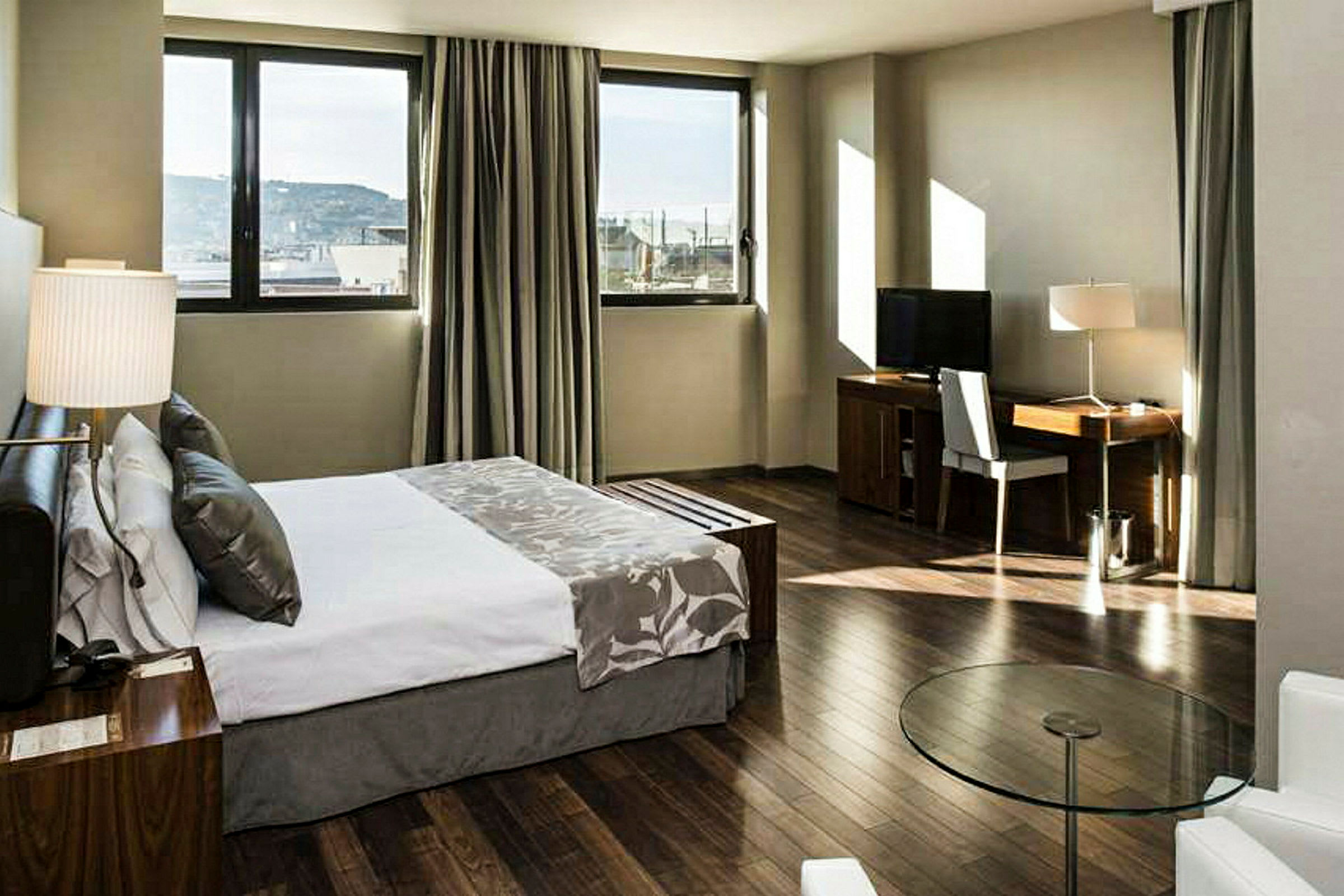 Ett hotellrum med en dubbelsäng, stora fönster, skrivbord med stol och TV, samt gardiner och trägolv. (AI-genererad text)