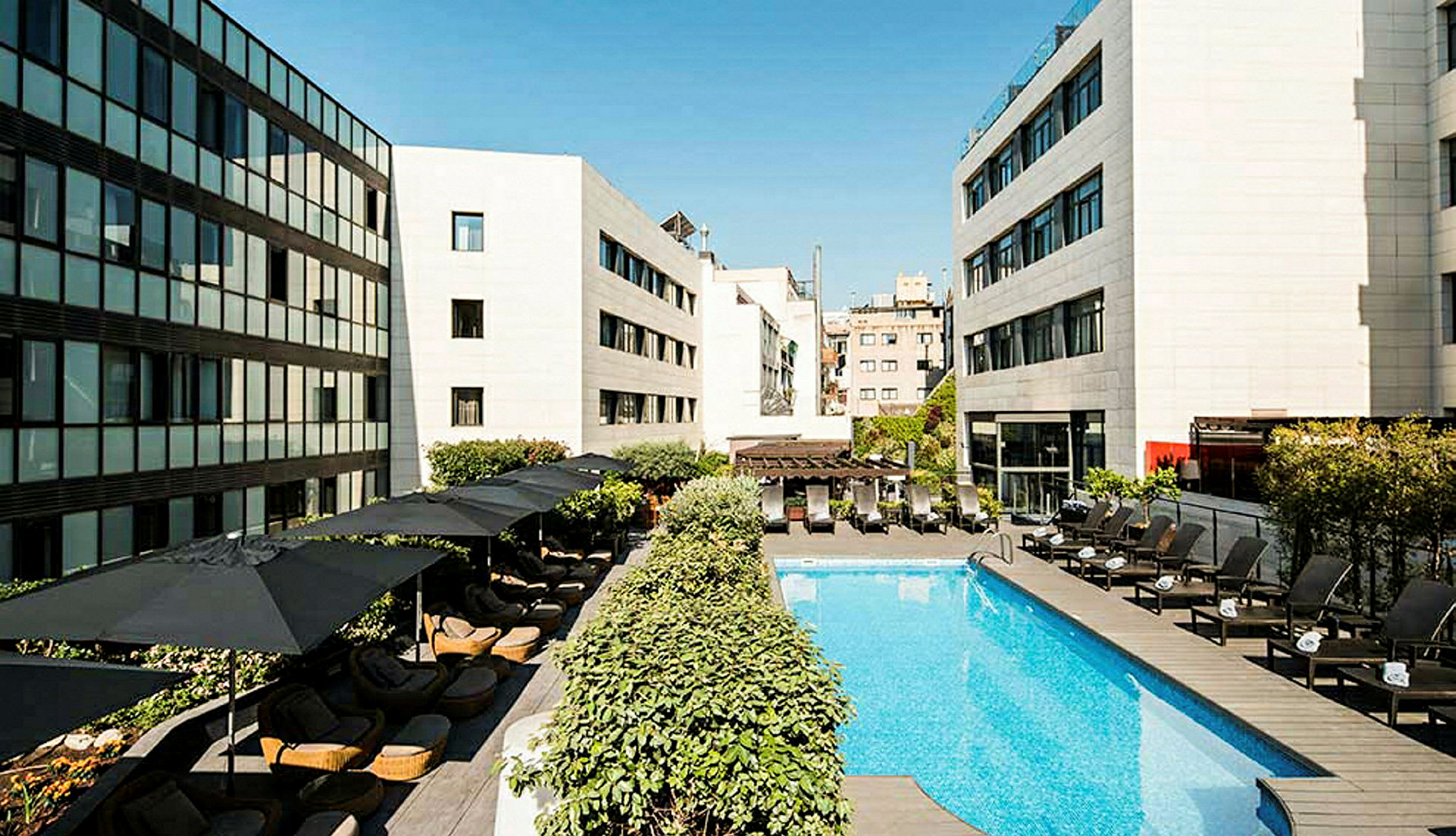 En hotellpool omgiven av liggstolar och parasoller, med moderna byggnader runtom. (AI-genererad text)