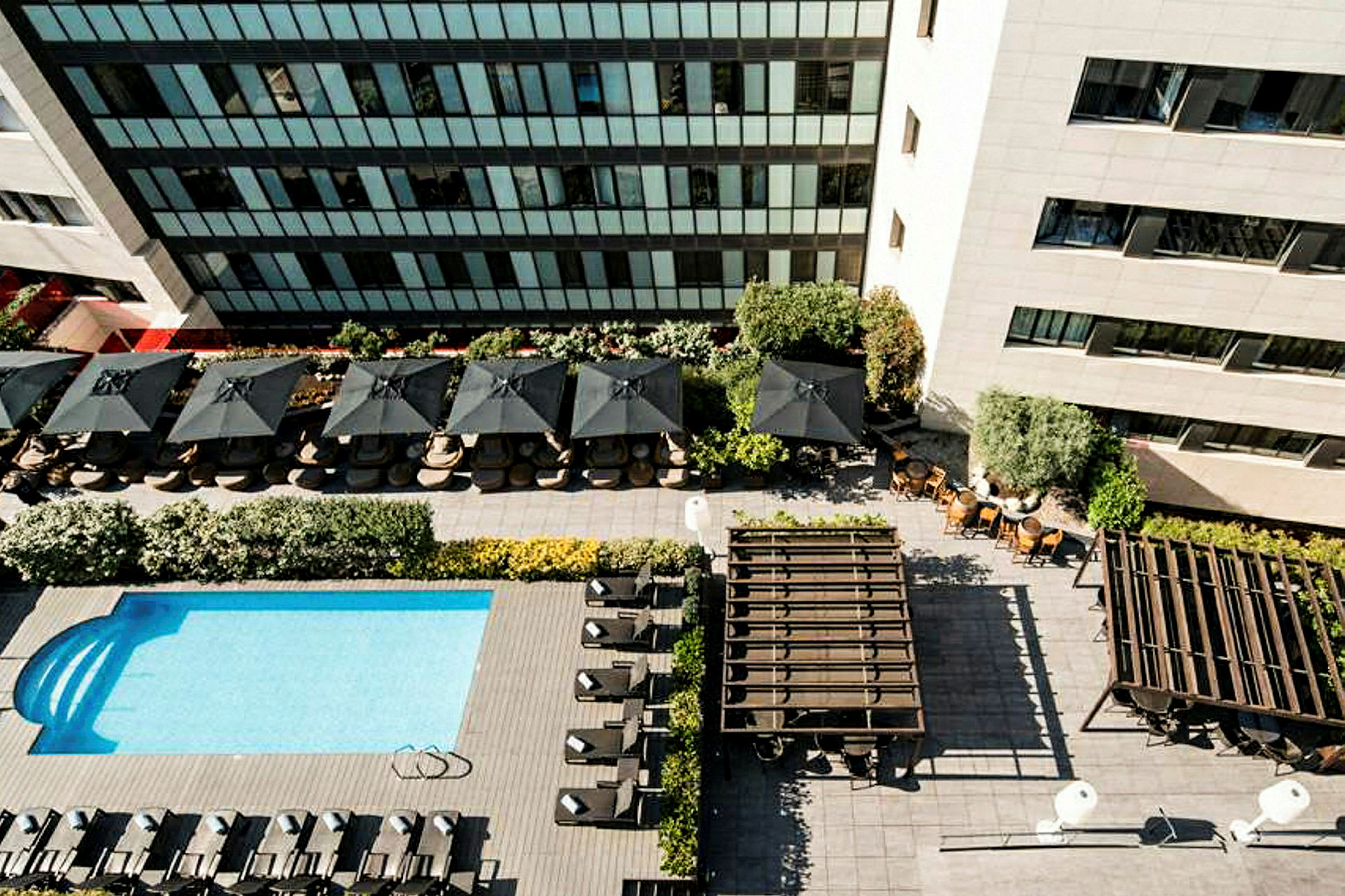 Flygbild över en pool med solstolar, parasoller och omgivande byggnader. (AI-genererad text)