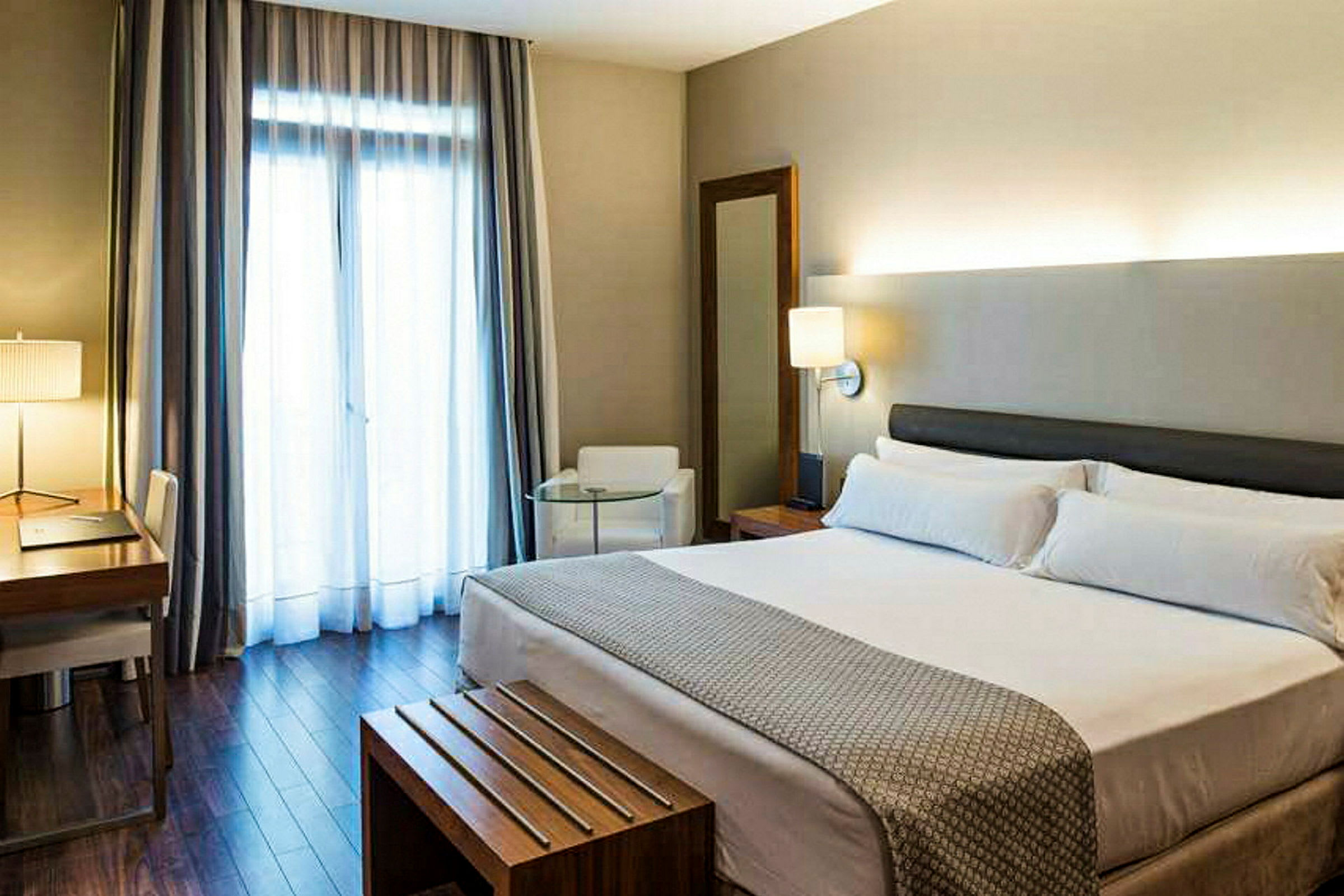 Ett hotellrum med en dubbelsäng, ett skrivbord och stol, en fåtölj, och ett stort fönster med gardiner. (AI-genererad text)