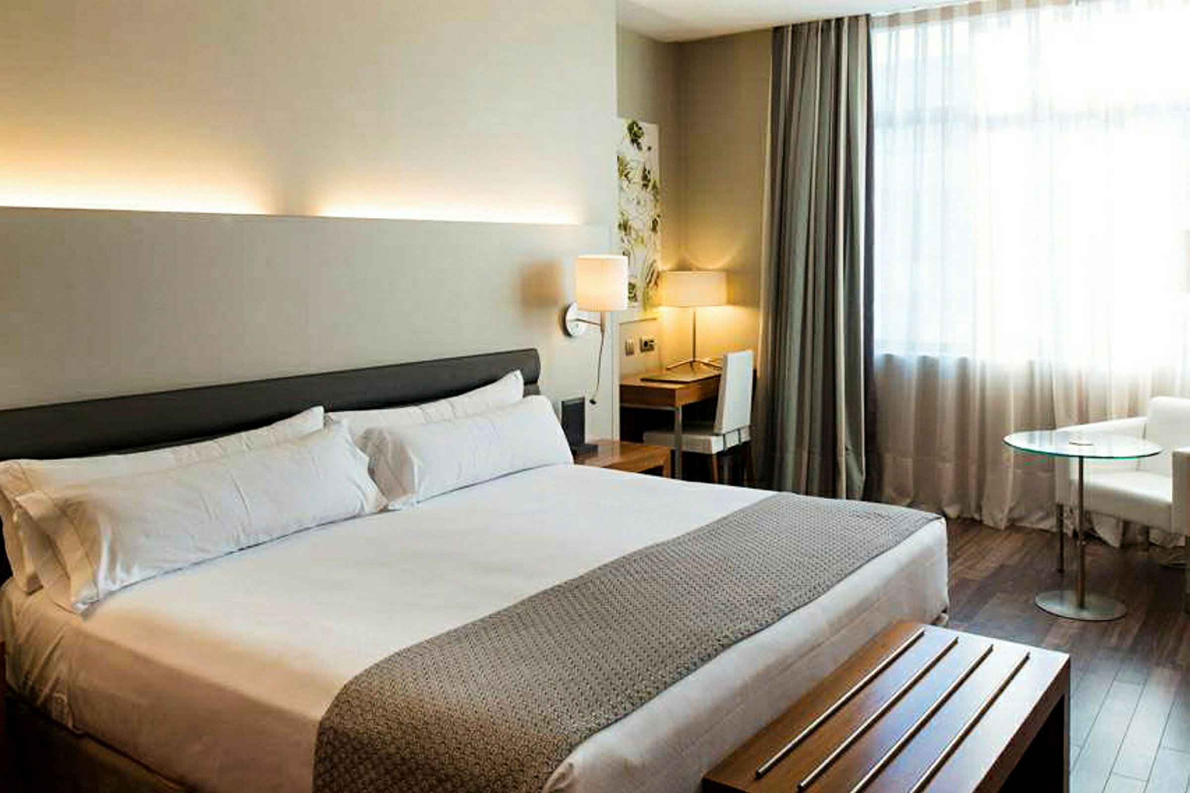 Ett hotellrum med en stor dubbelsäng, ett skrivbord med stol och lampa samt en fåtölj och ett glasbord vid fönstret. (AI-genererad text)