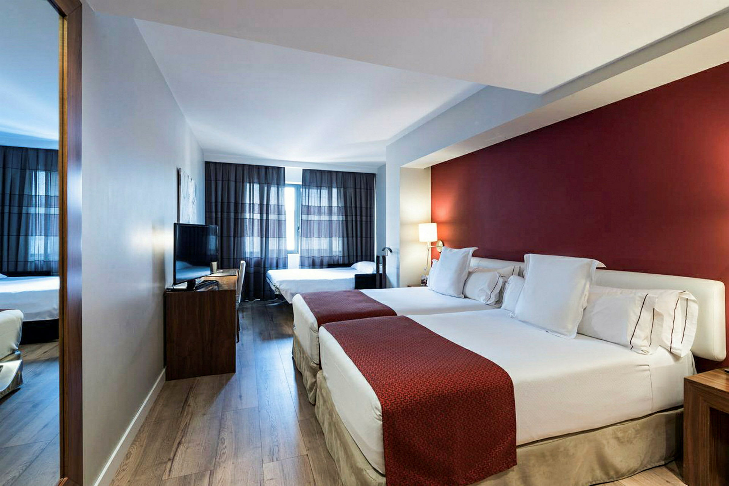 Ett hotellrum med två stora sängar, röda dekorativa överkast, en röd väggfärg, och en stor fönsteryta med mörka gardiner. (AI-genererad text)