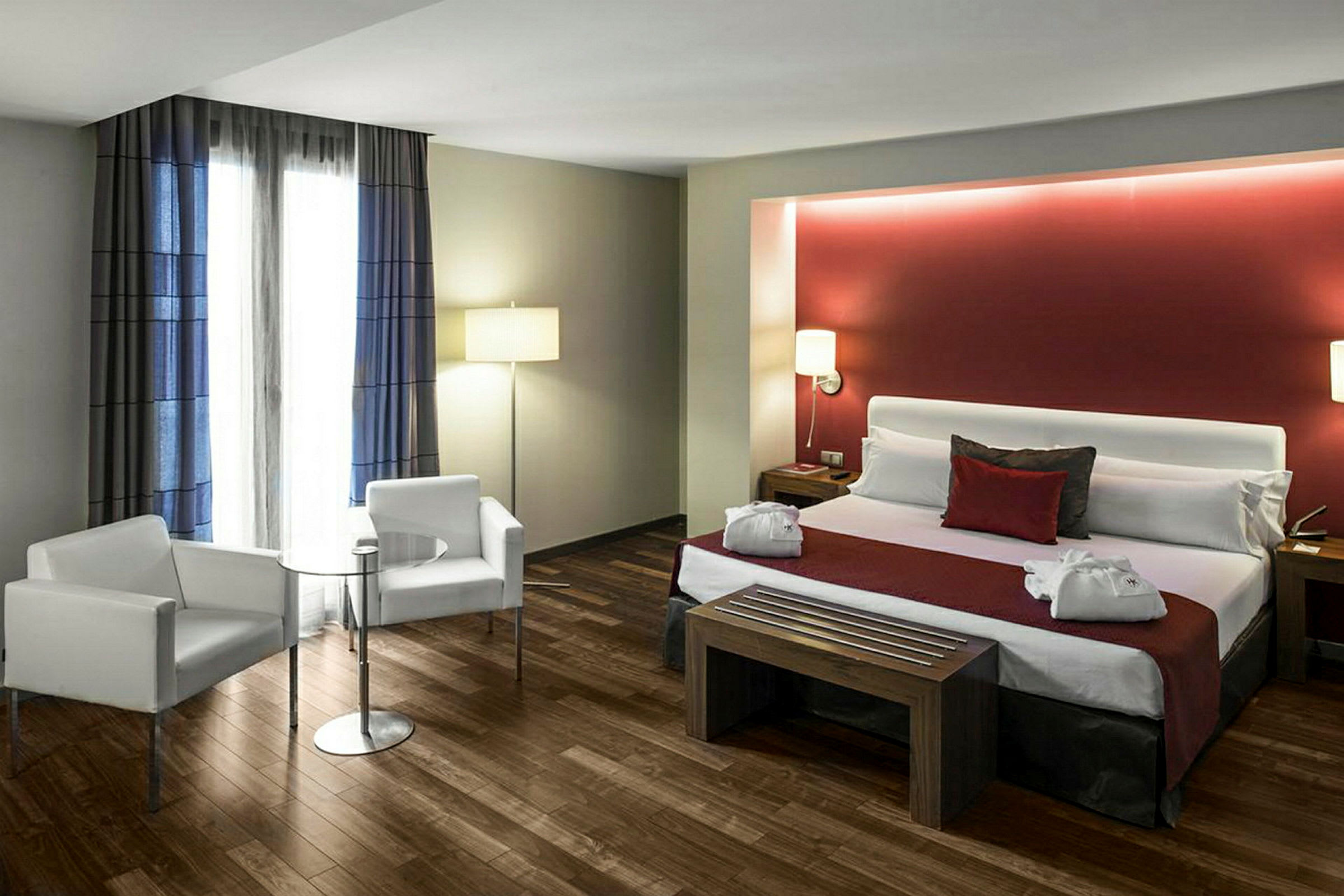 Ett hotellrum med en dubbelsäng och prydnadskuddar, flankerad av sängbord med lampor. Det finns två vita fåtöljer och ett litet bord framför ett fönster med gardiner. (AI-genererad text)