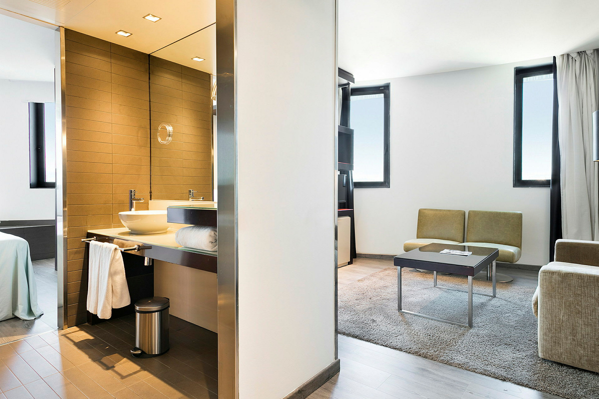 Ett modernt hotellrum som visar badrum med tvättställ och spegel till vänster, sovrummet med säng längre bak, samt vardagsrum med soffor, bord och matta till höger. (AI-genererad text)