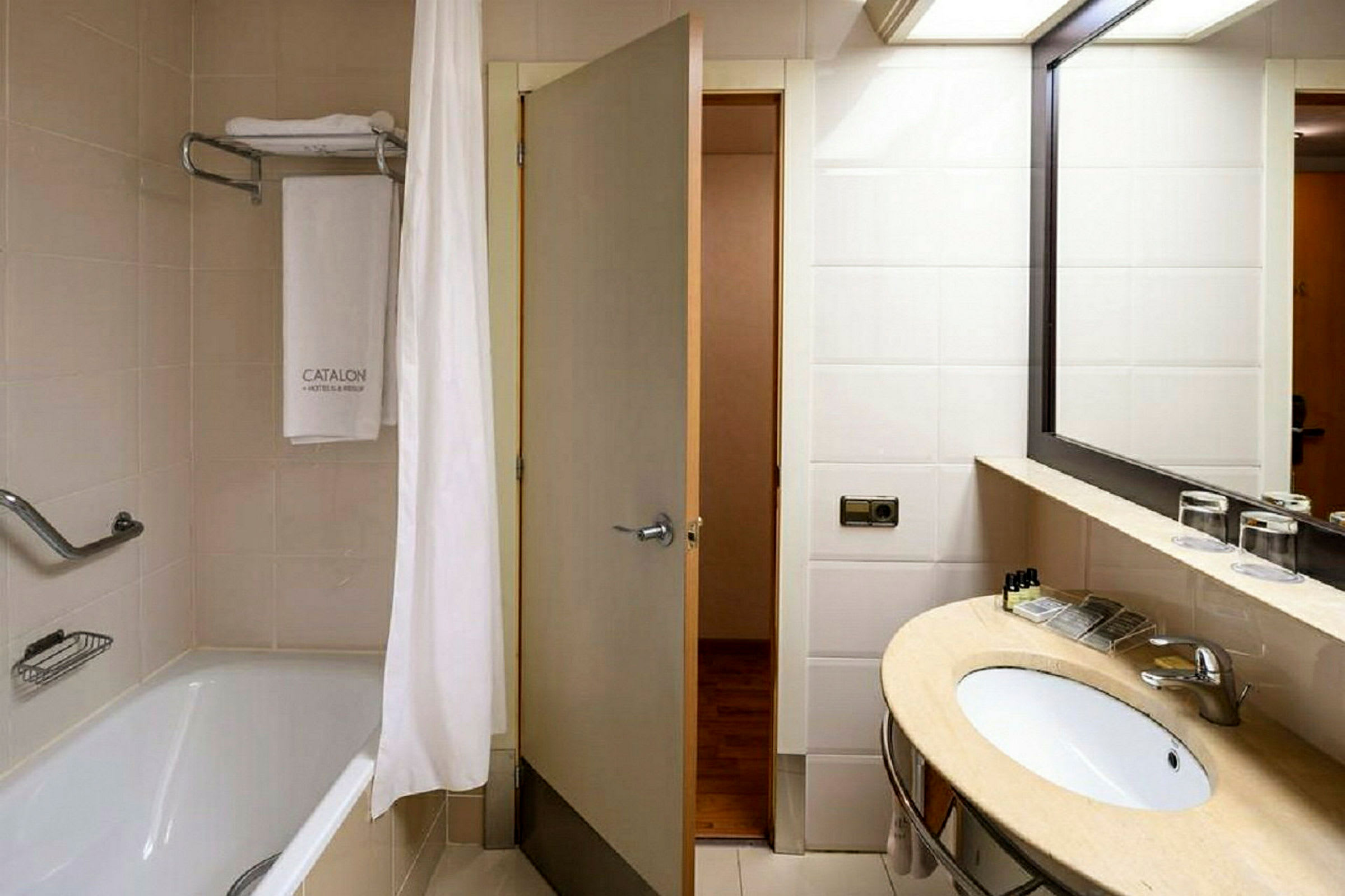 Hotellbadrum med ett badkar med duschdraperi, en handduk hängande på en stång och ett tvättställ med spegel. (AI-genererad text)
