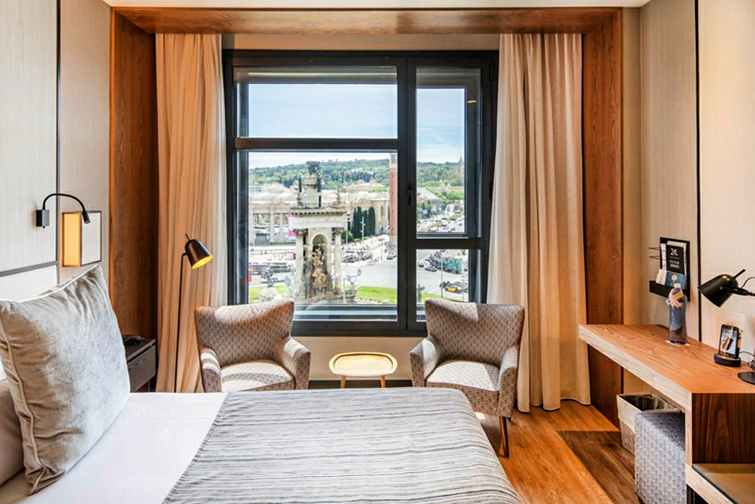 Ett hotellrum med en stor säng, två fåtöljer framför ett fönster med en utsikt över en stad, samt ett skrivbord på sidan. (AI-genererad text)