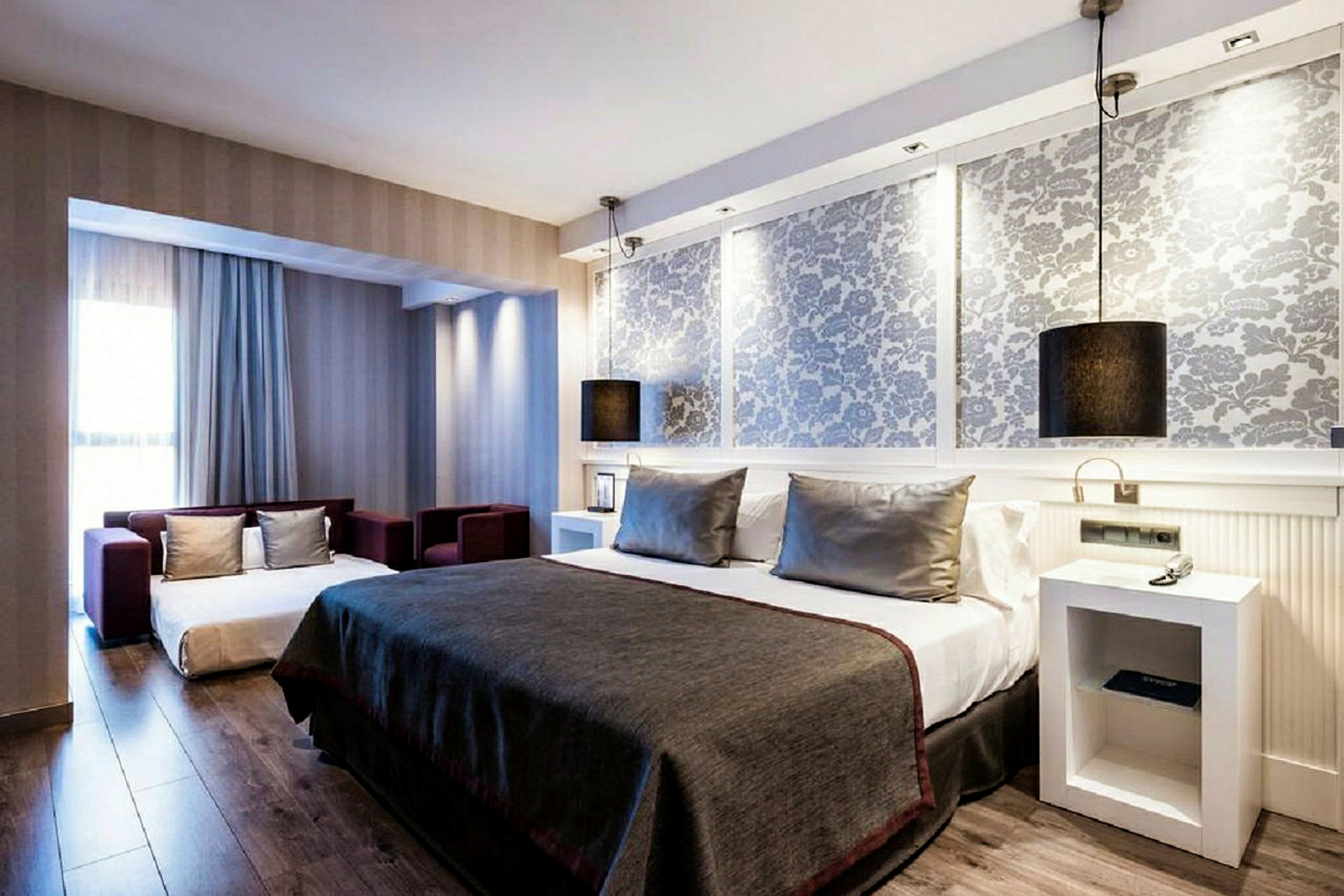 Ett modernt hotellrum med grå inredning, en stor säng med grå överkast och kuddar, samt en liten sittgrupp framför ett stort fönster. (AI-genererad text)