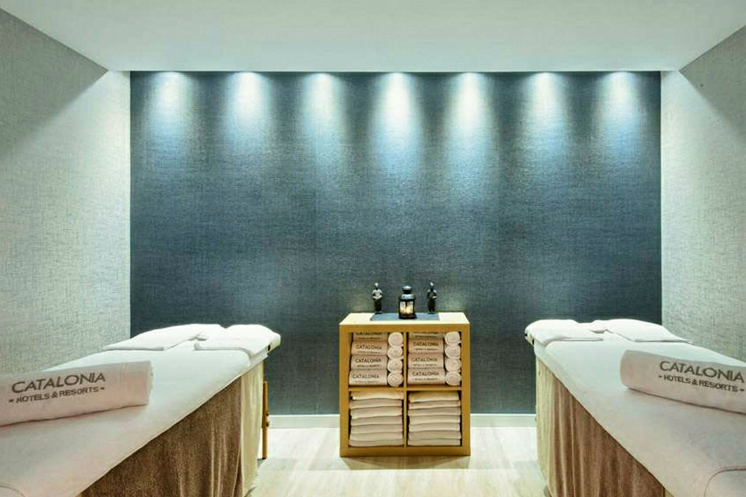 Ett spa-rum med två massagebänkar, prydliga handdukar och dekorativa föremål på en hylla mellan bänkarna. (AI-genererad text)