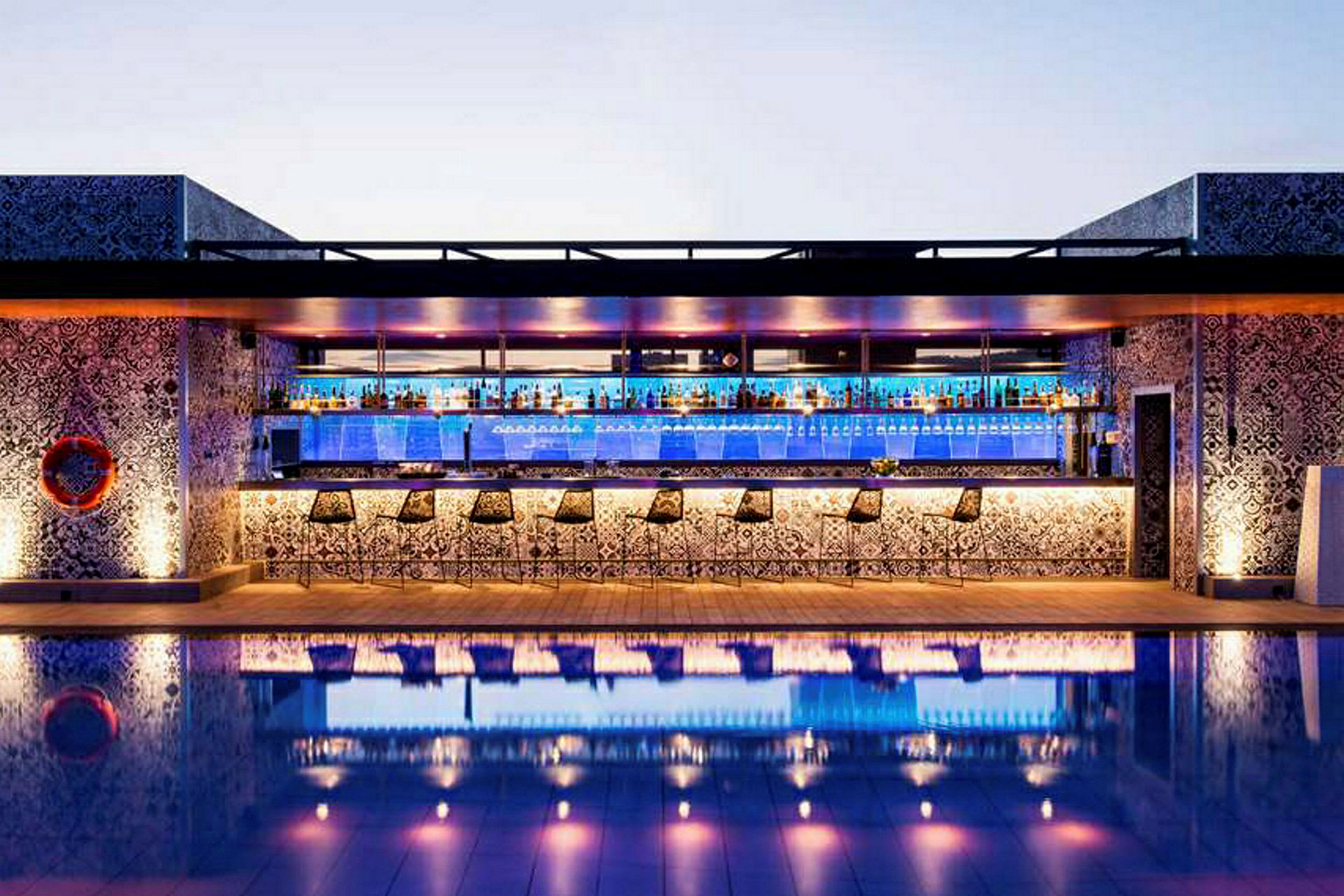 En utomhusbar med barstolar framför och dekorativa mönster på väggarna, reflekteras i en pool. (AI-genererad text)