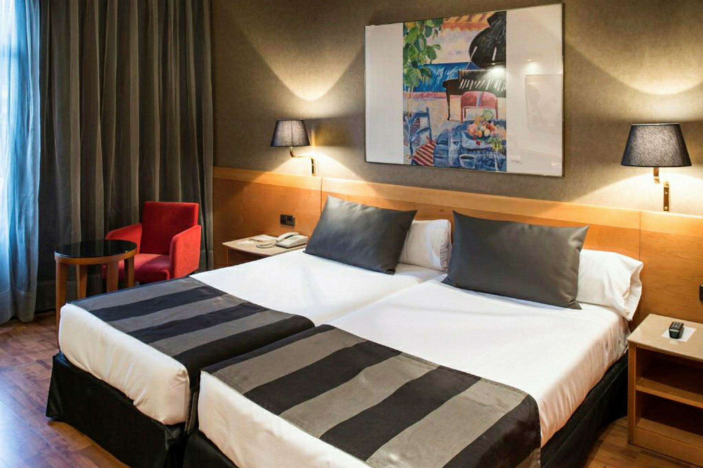 Ett hotellrum med två enkelsängar, varav båda är bäddade med svartvita överkast. På väggen hänger en färgglad tavla, och rummet har även ett litet bord, en röd fåtölj och två vägglampor. (AI-genererad text)