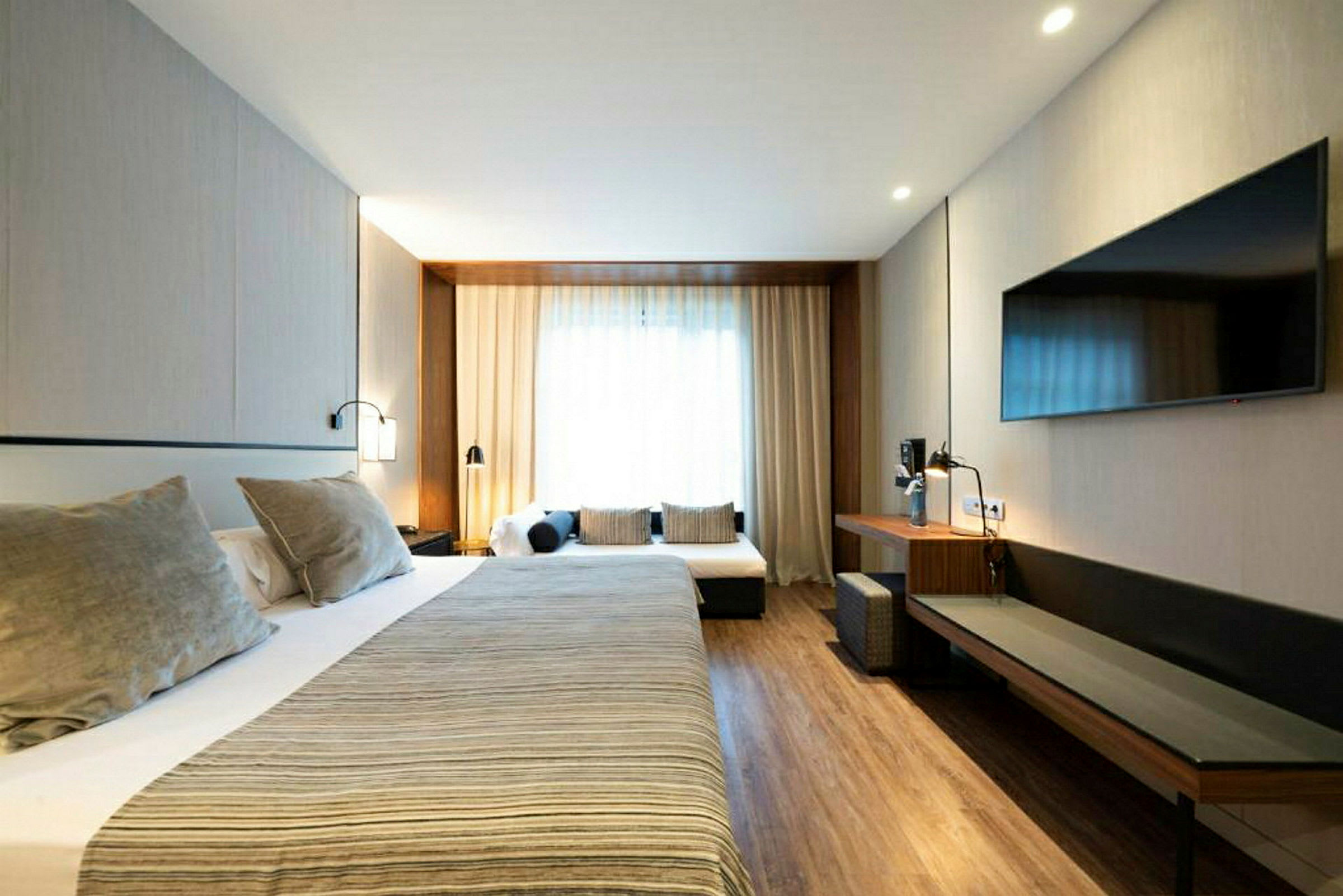 Ett hotellrum med en stor säng, därtill hörande sängbord och lampor samt en sittgrupp med soffkuddar. Rummet har trägolv och en väggmonterad TV. (AI-genererad text)