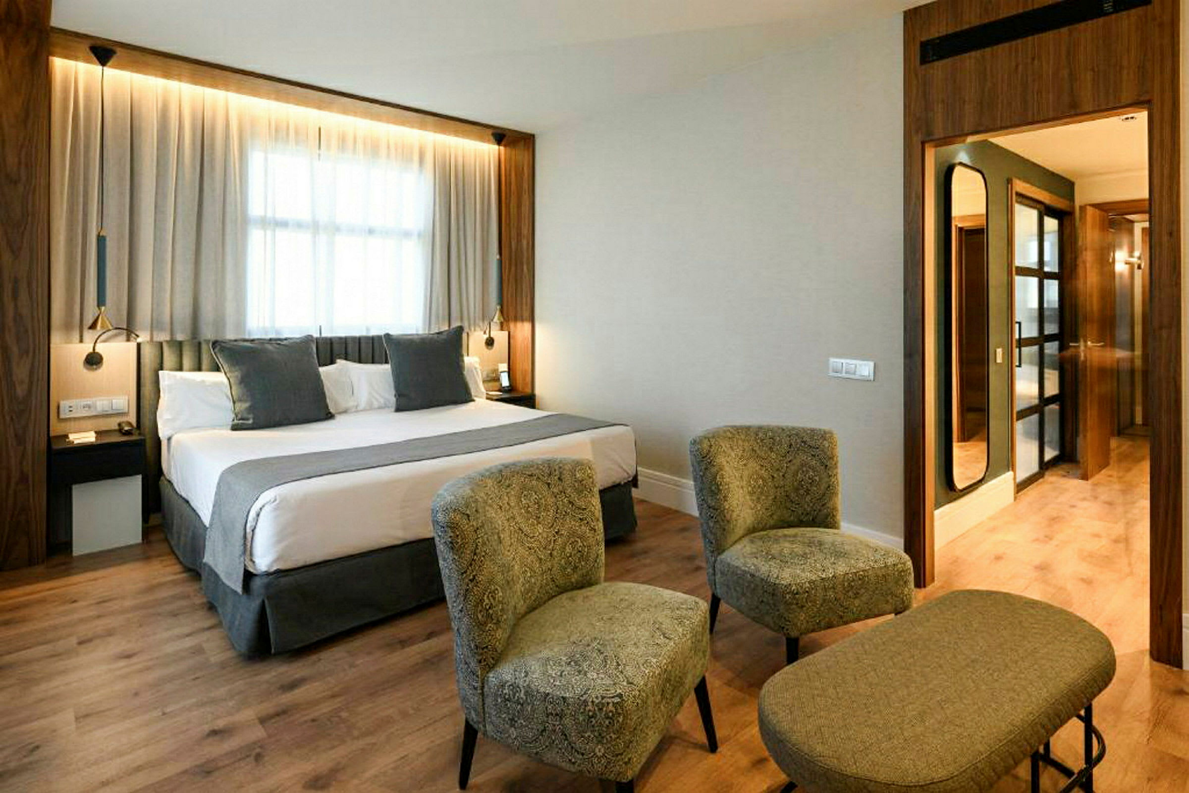 Ett hotellrum med en dubbelsäng, två stolar och ett litet bord framför. Rummet har en ljus inredning och ett stort fönster med gardiner. (AI-genererad text)