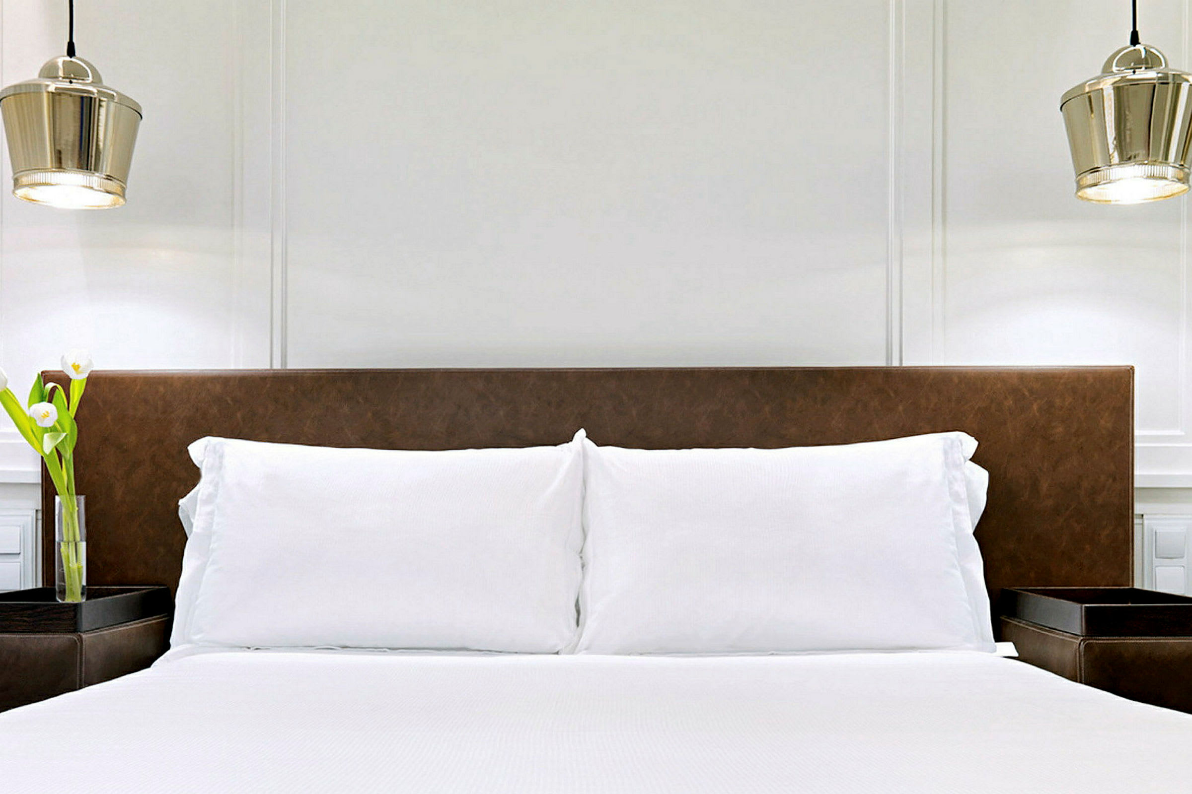 Ett modernt hotellrum med en dubbelsäng, vita sängkläder och brunt sänggavel. På ena sidan av sängen står en vas med blommor. (AI-genererad text)