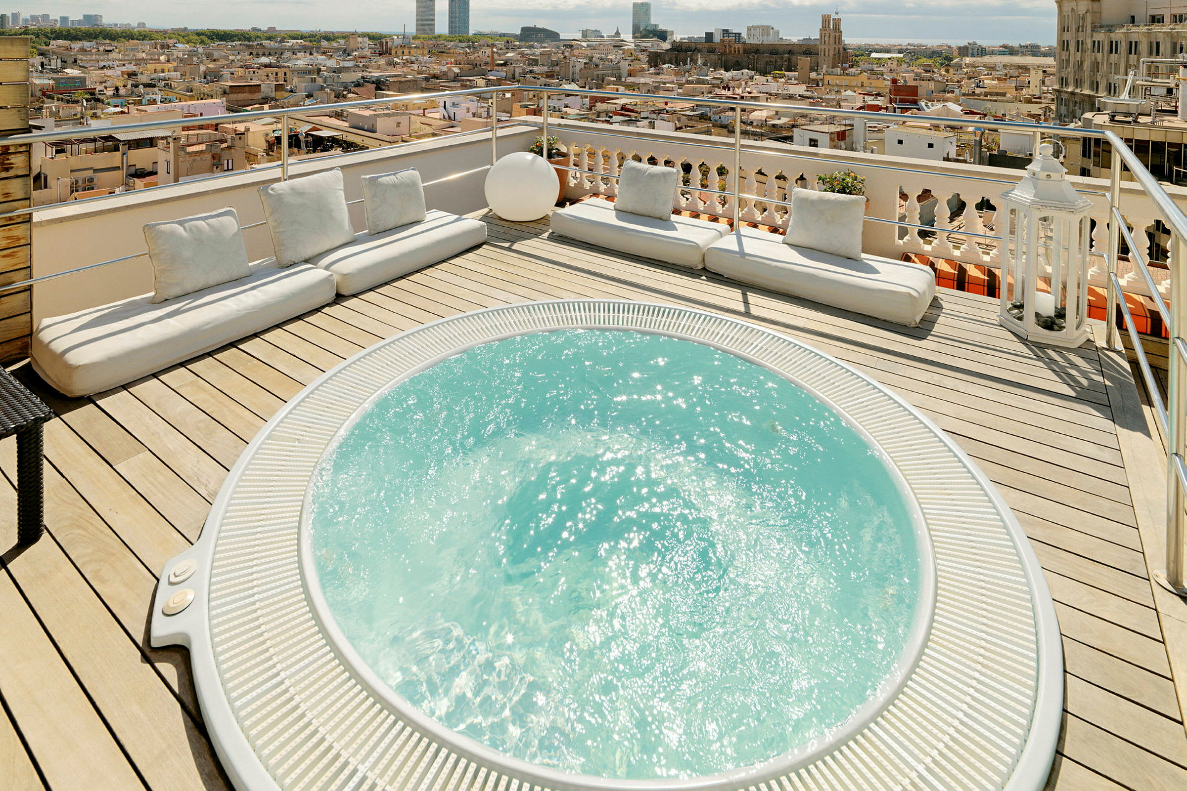En terrass med en jacuzzi och flera vita sittdynor, med utsikt över en stad. (AI-genererad text)