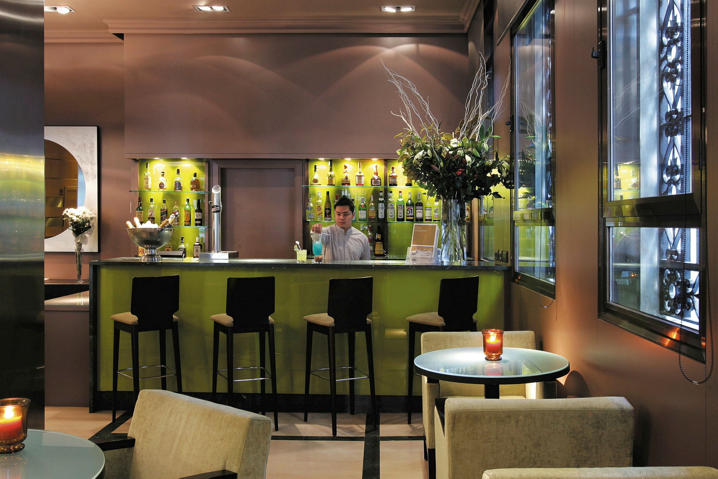 En modern bar med grön belysning och flera hyllor fyllda med flaskor. Framför baren står höga stolar och runt om finns bord och fåtöljer. (AI-genererad text)
