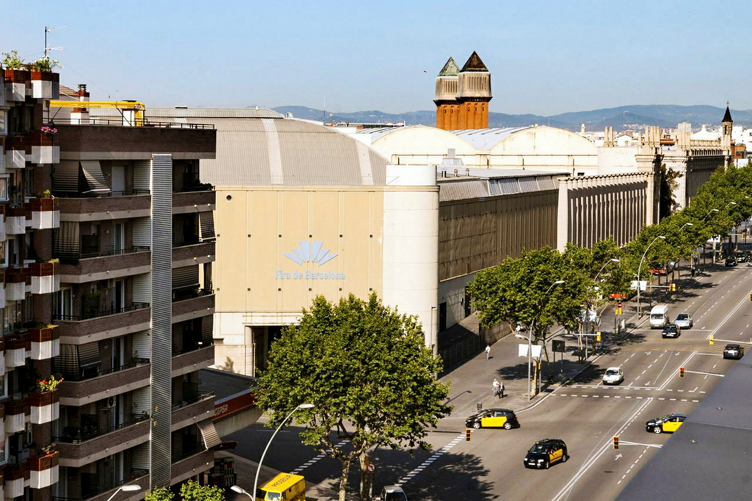 En gata med träd och fordon framför stora byggnader, inklusive en med skylten 'Fira de Barcelona'. (AI-genererad text)