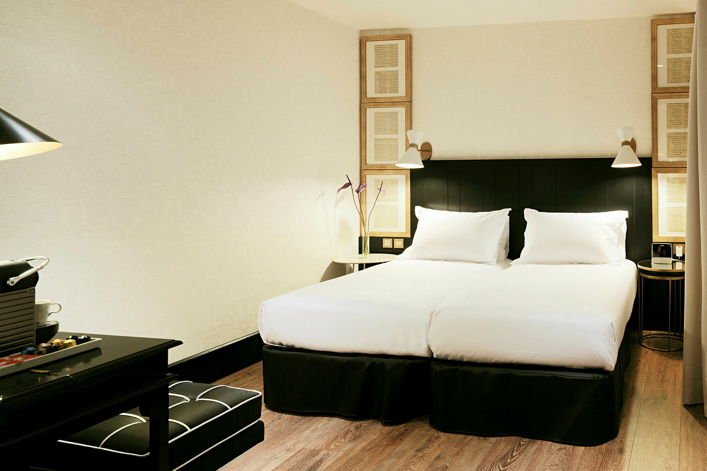 Ett hotellrum med två enkelsängar, vit sängduk och matchande kuddar. Inredningen inkluderar mörka möbler och dekorativa detaljer. (AI-genererad text)