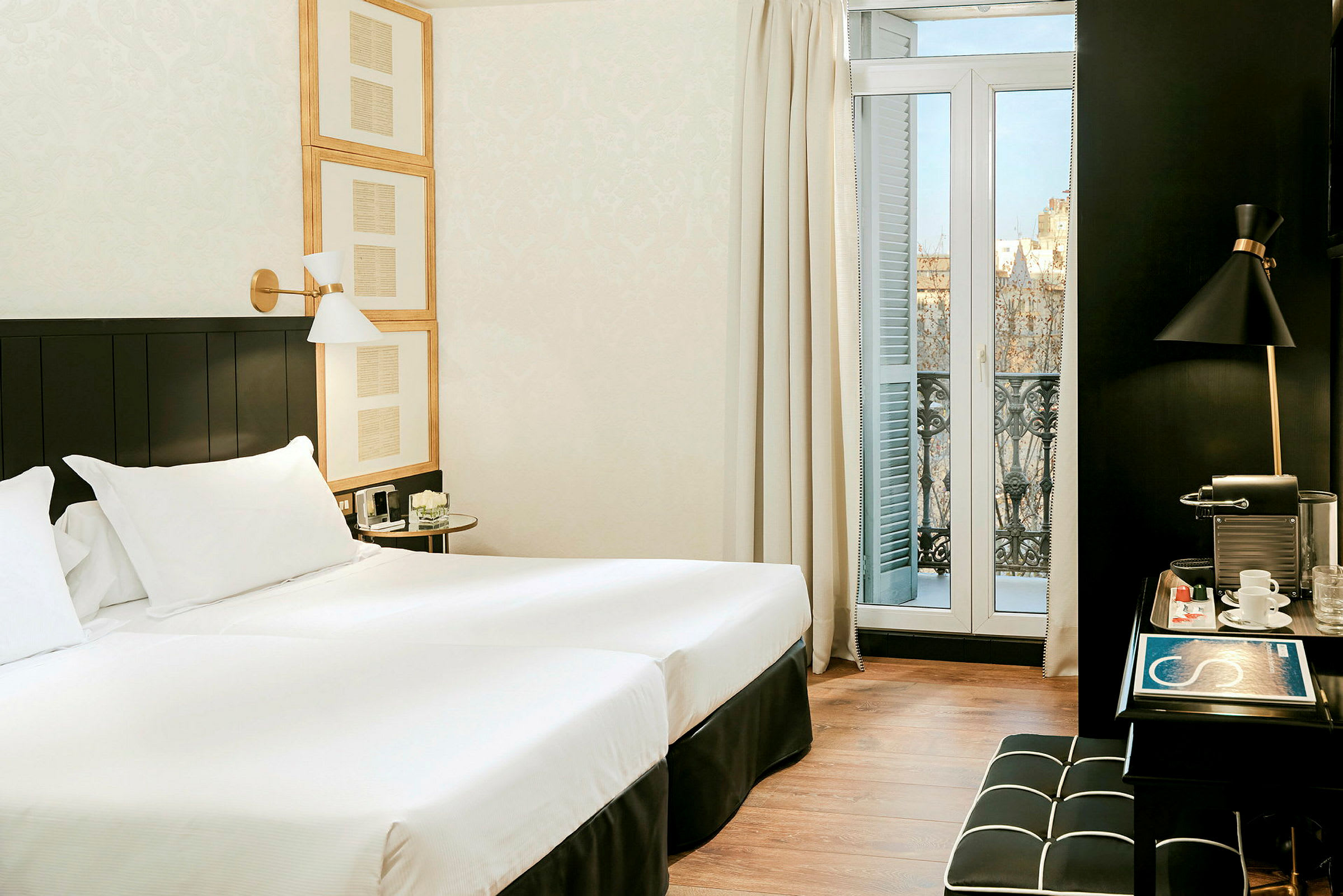 Ett hotellrum med två enkelsängar, vita lakan och mörkt träinredning. Ett skrivbord med kaffemaskin och en liten balkong med utsikt finns. (AI-genererad text)
