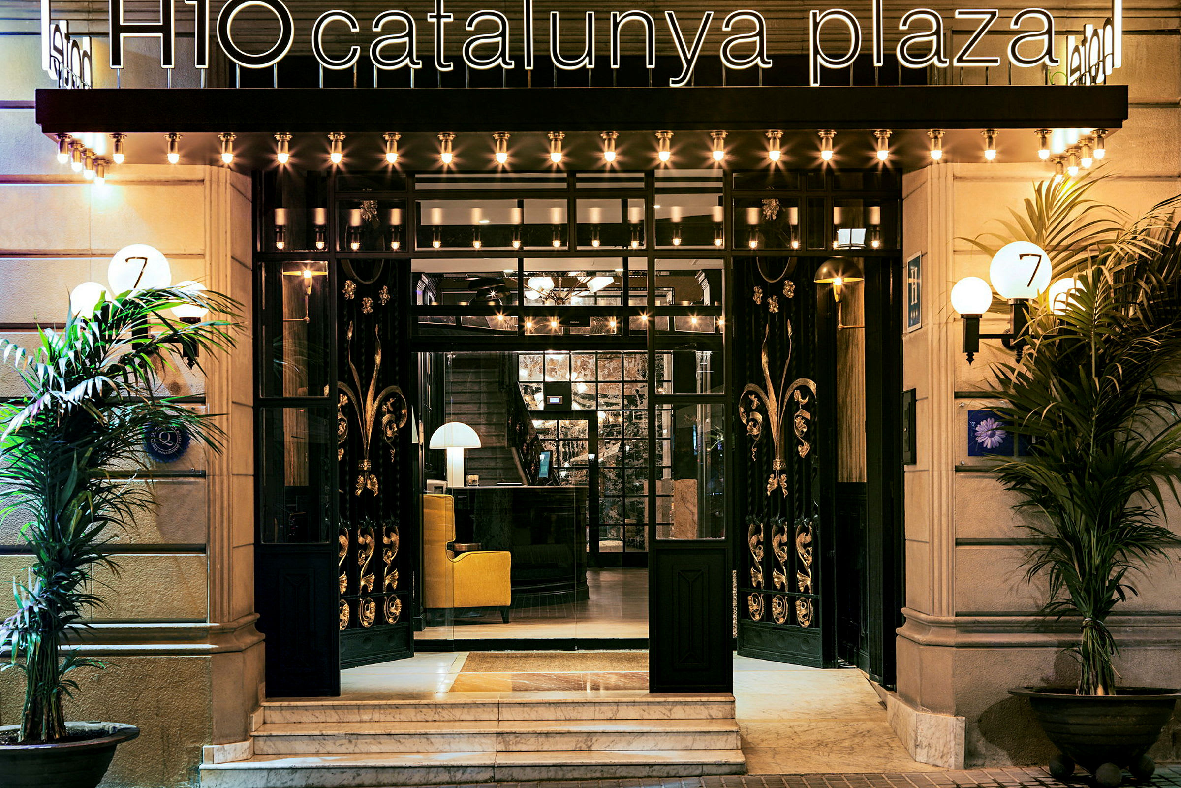 Entrén till ett hotell med en skylt "H10 Catalunya Plaza" och prydnader runt dörrarna. (AI-genererad text)