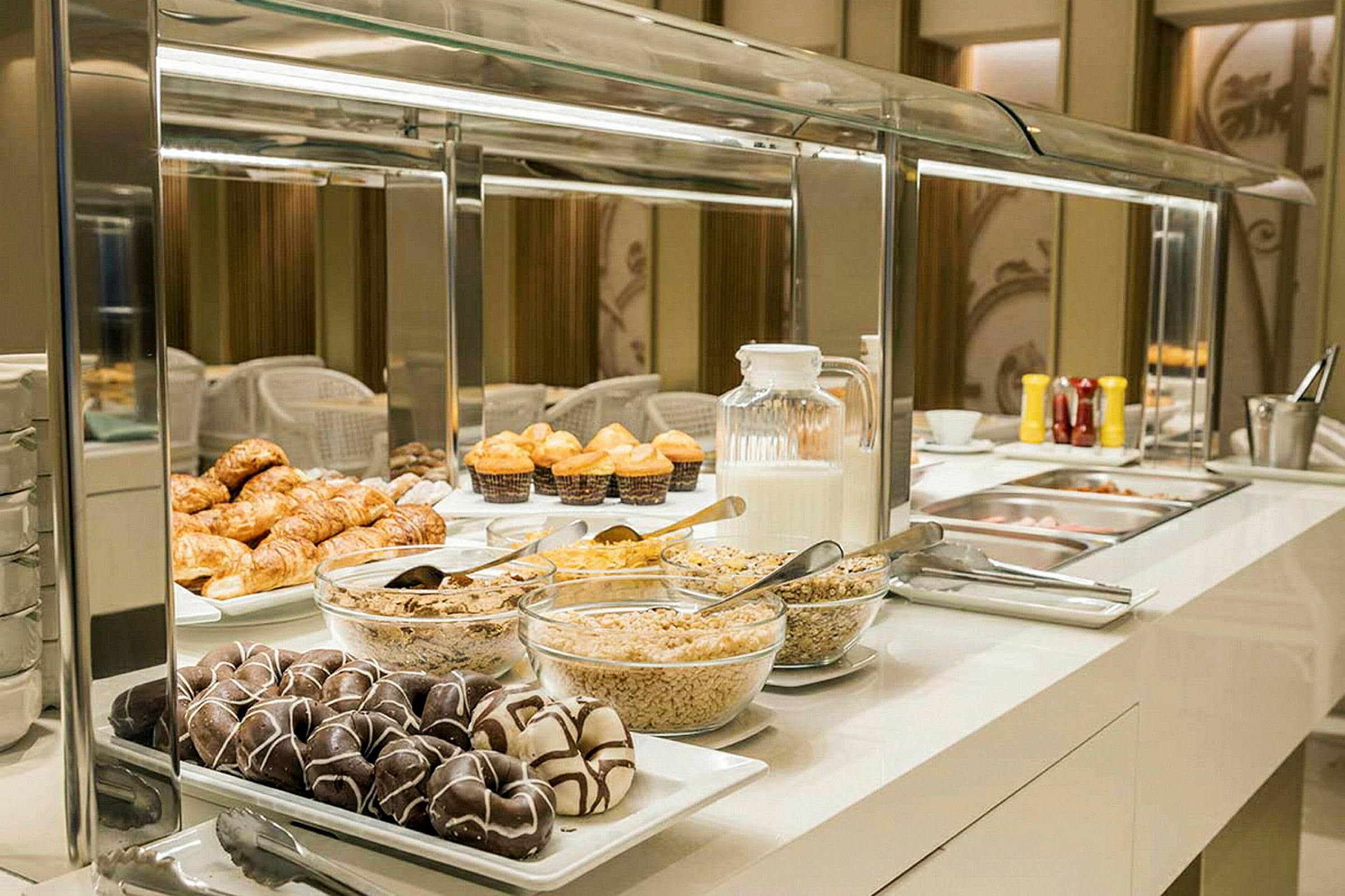 Frukostbuffé med olika bakverk, flingor och tillbehör samt en kanna mjölk på ett upplyst serveringsbord. (AI-genererad text)
