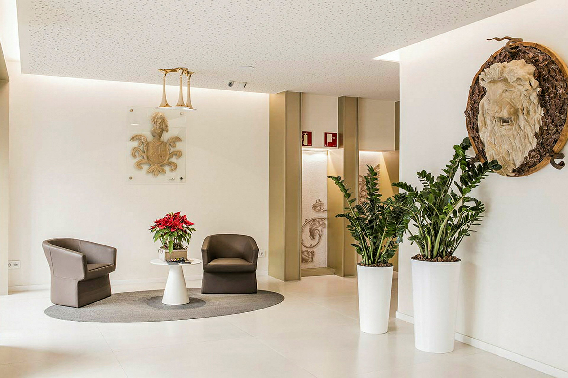 Ett elegant hotellutseende lobbyområde med två bruna fåtöljer och ett vitt bord med en blomvas på, omgiven av grönska och dekorerade väggdetaljer. (AI-genererad text)