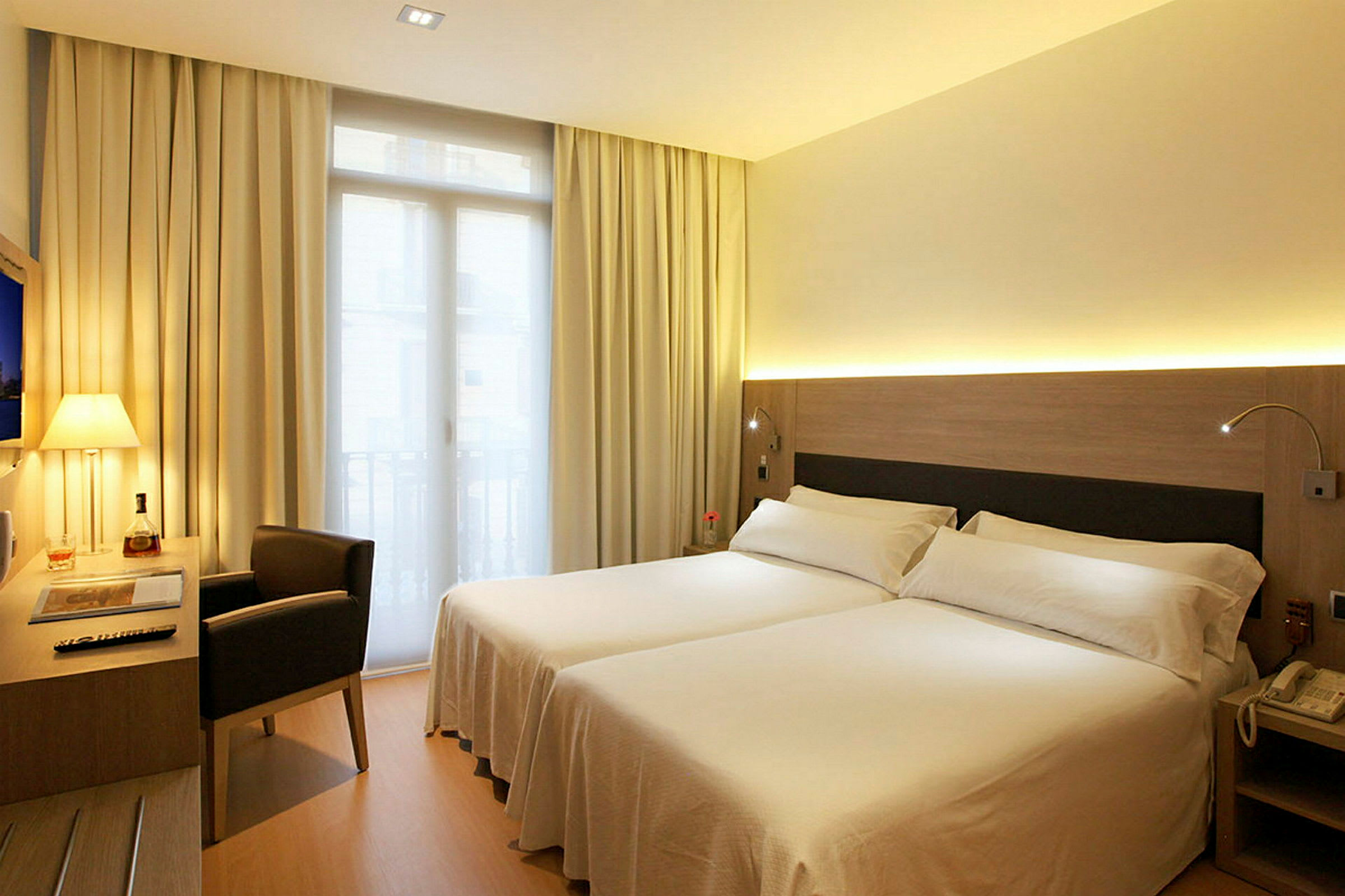 Ett hotellrum med två enkelsängar, ett skrivbord och en stol vid sidan av en fönsterdörr. (AI-genererad text)